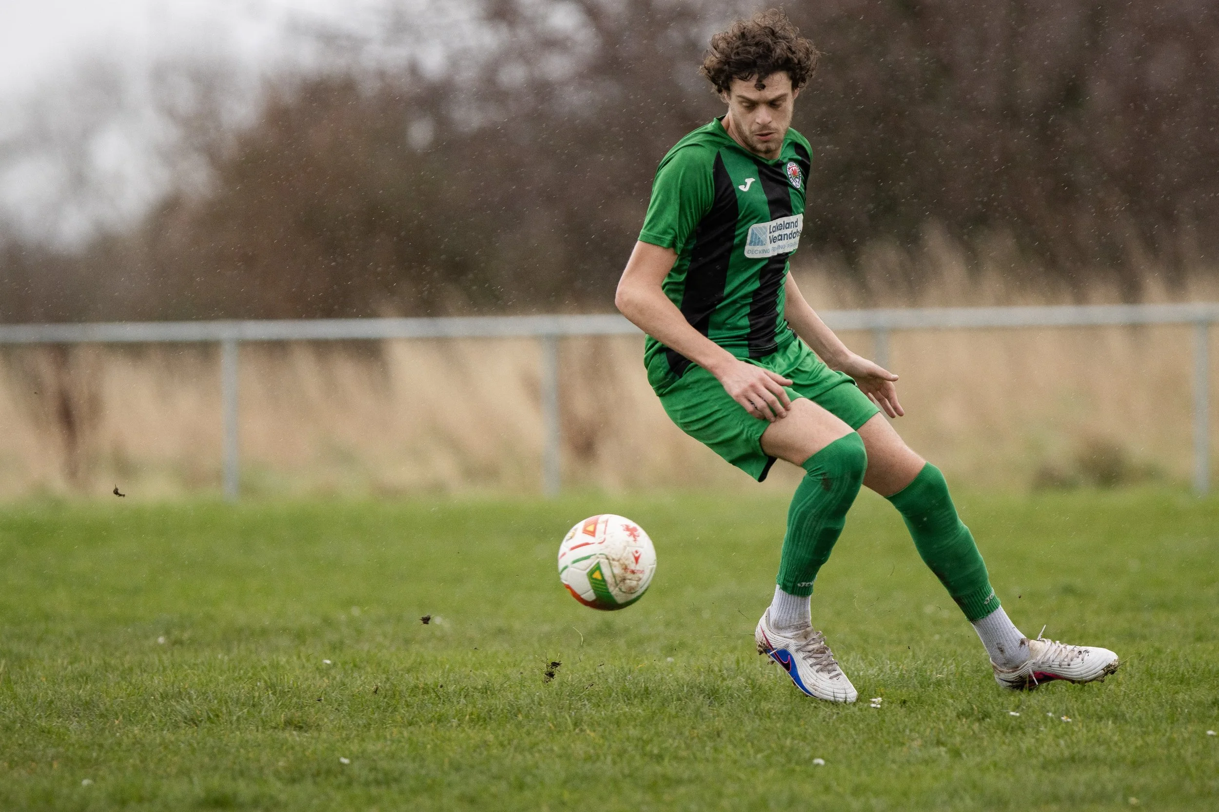 A male soccer player in a green uniform kicks a soccer ball on a grassy field during rainy conditions.