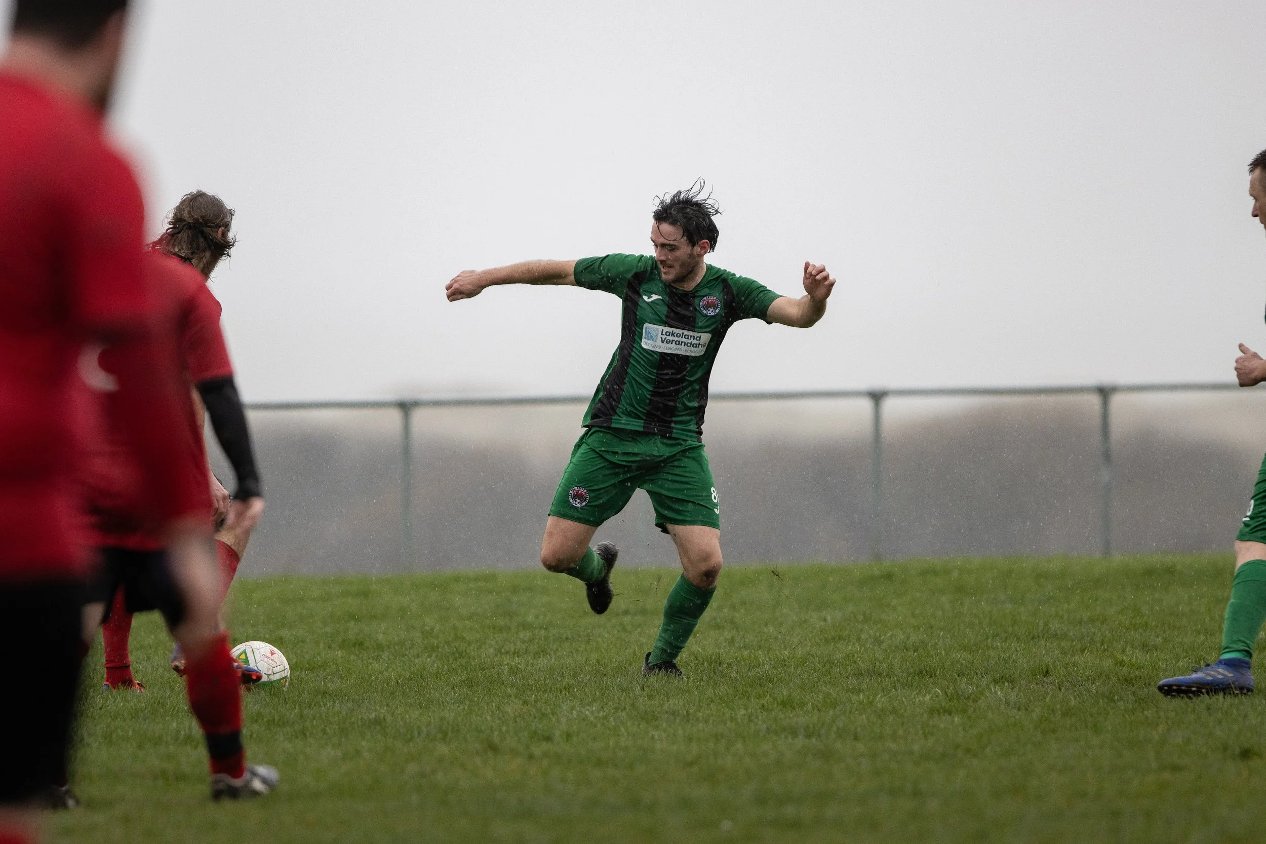 Soccer players playing on a rainy field, with one player in green kicking a ball while others watch.