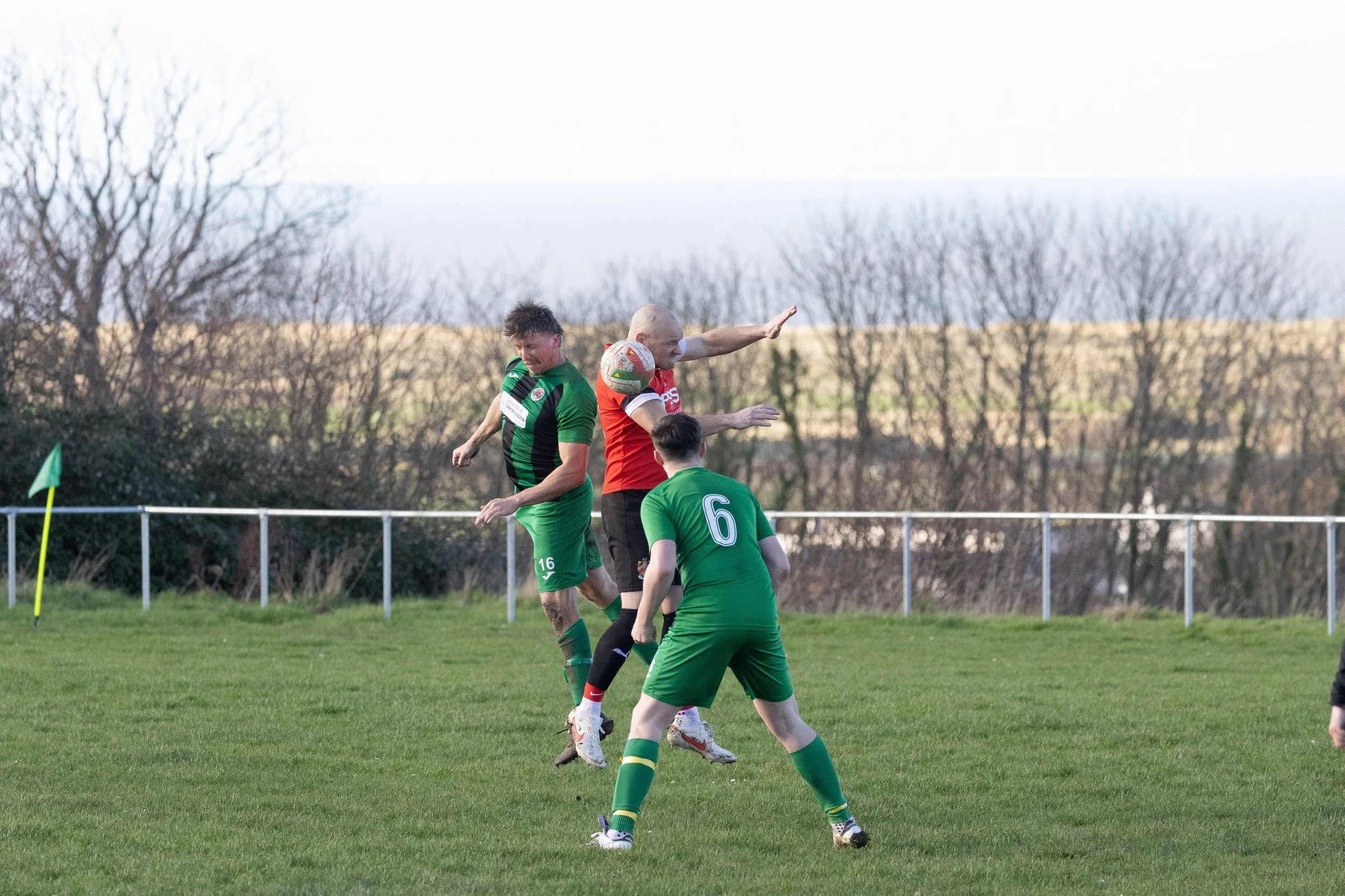 Three soccer players and a referee jump to head the ball during a game on a grassy field, with trees and a fence in the background.