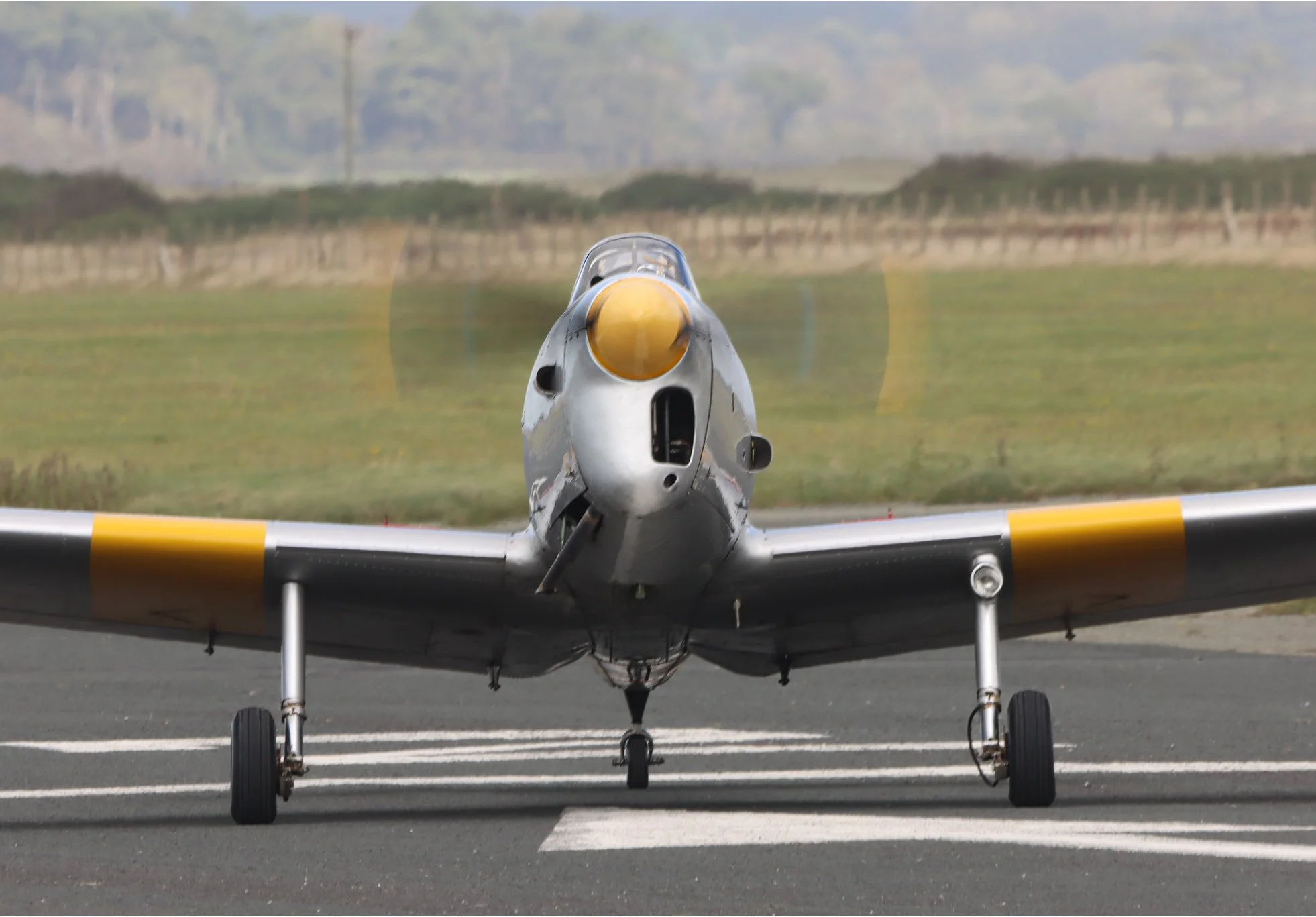 A military fighter jet taxiing on the runway with farmland and trees in the background.