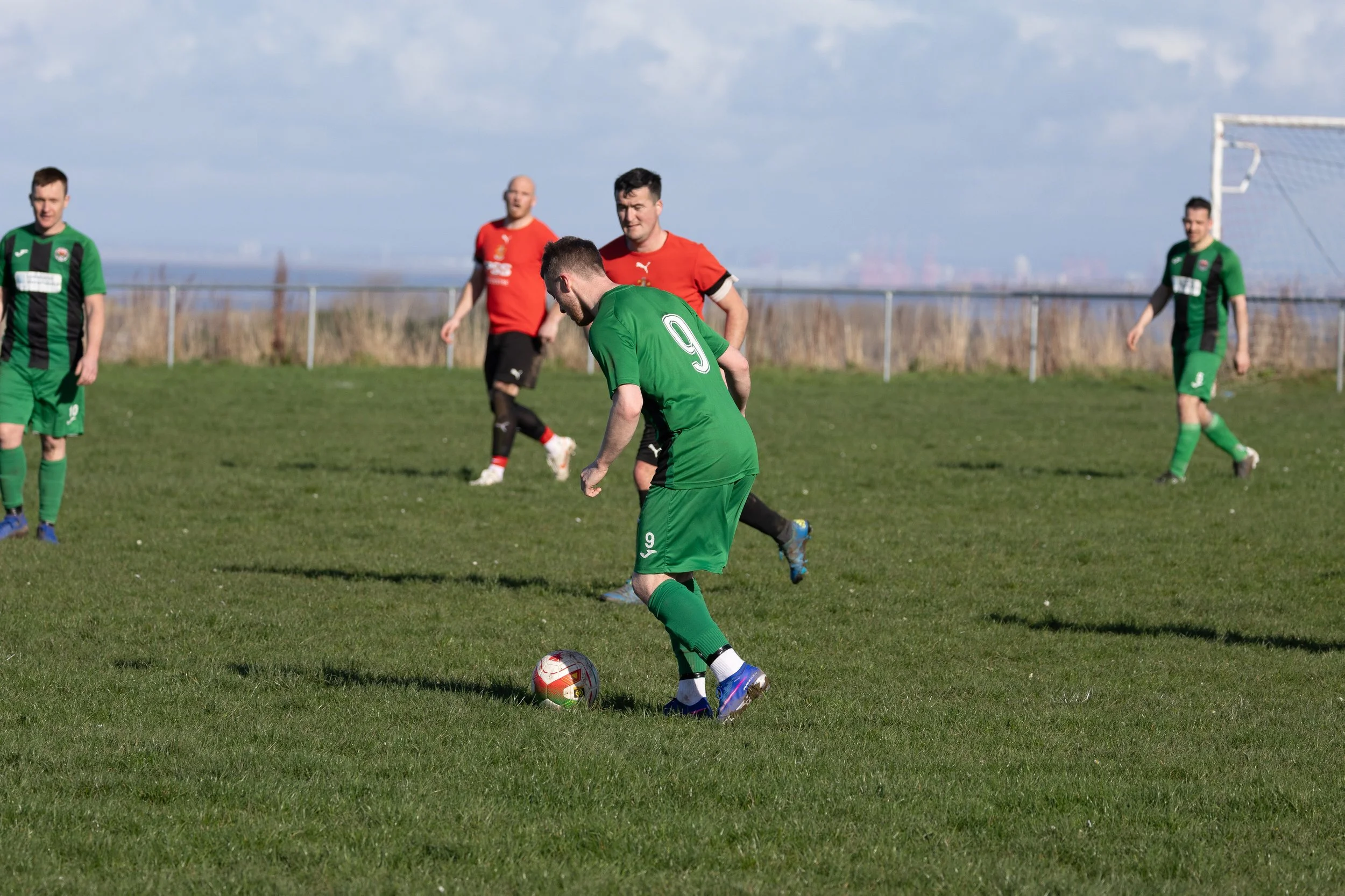 Soccer players on green field with two in red and three in green uniforms during a game, one in green about to kick the ball.