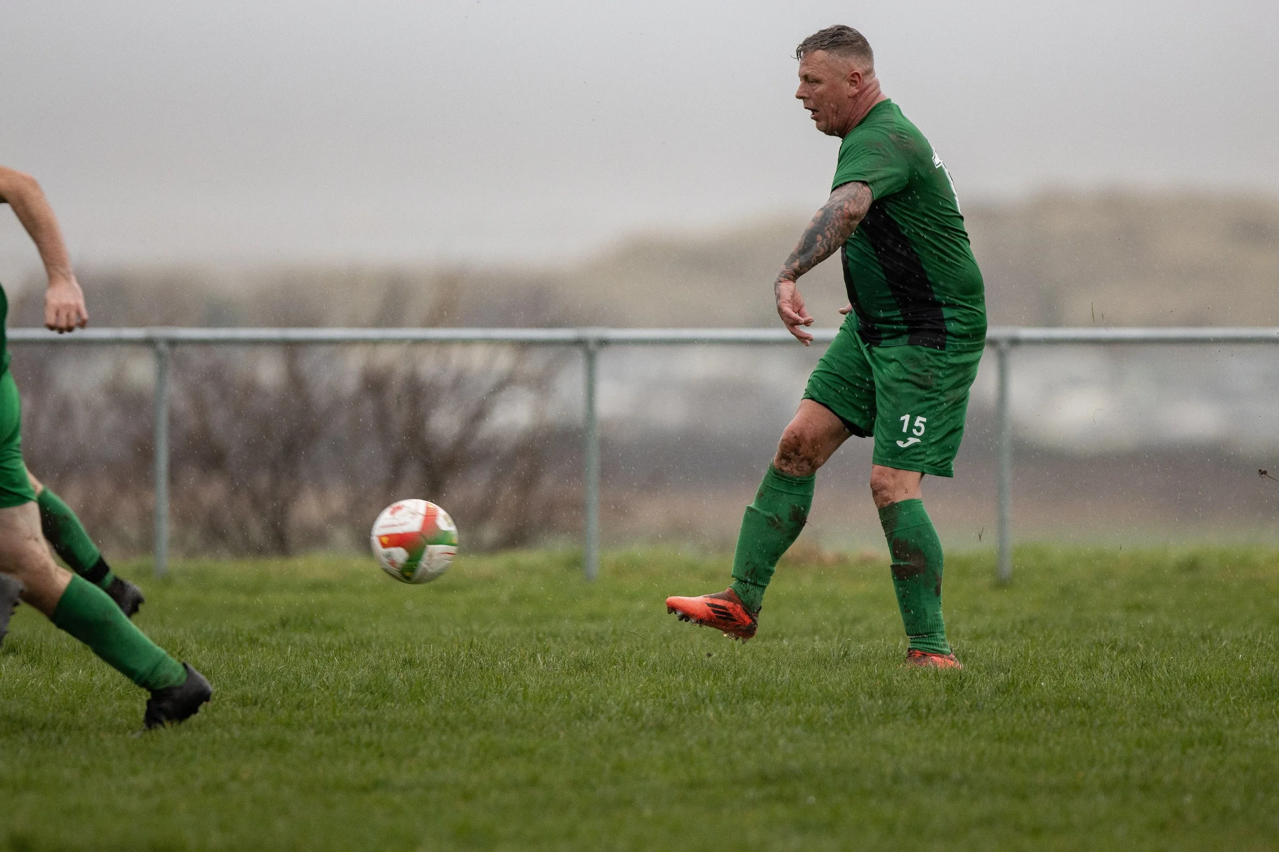 A soccer player wearing a green uniform with the number 15, kicking a soccer ball on a grassy field in rainy weather.