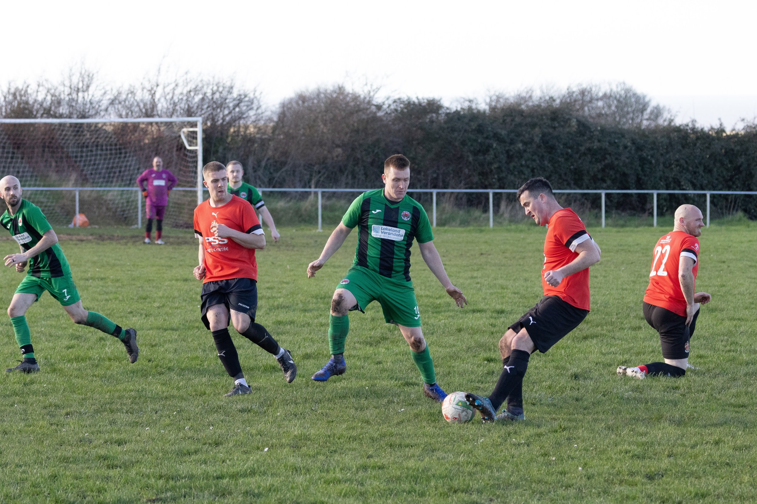 Soccer players in green and orange jerseys competing for the ball on a grassy field, with a goal and a goalkeeper in the background, during late afternoon or early evening.