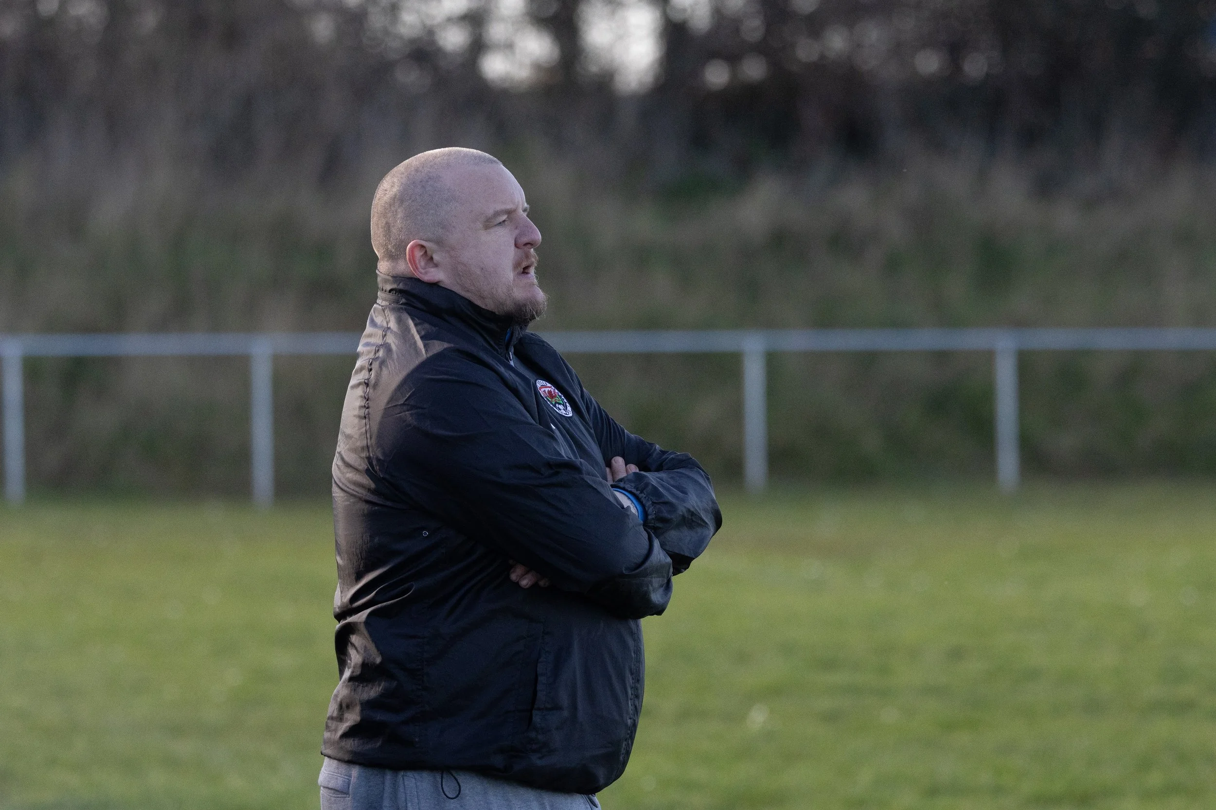 A man stands on a grassy field with his arms crossed, wearing a black jacket with a badge on it, looking thoughtfully into the distance. Background features a blurred fence and trees.