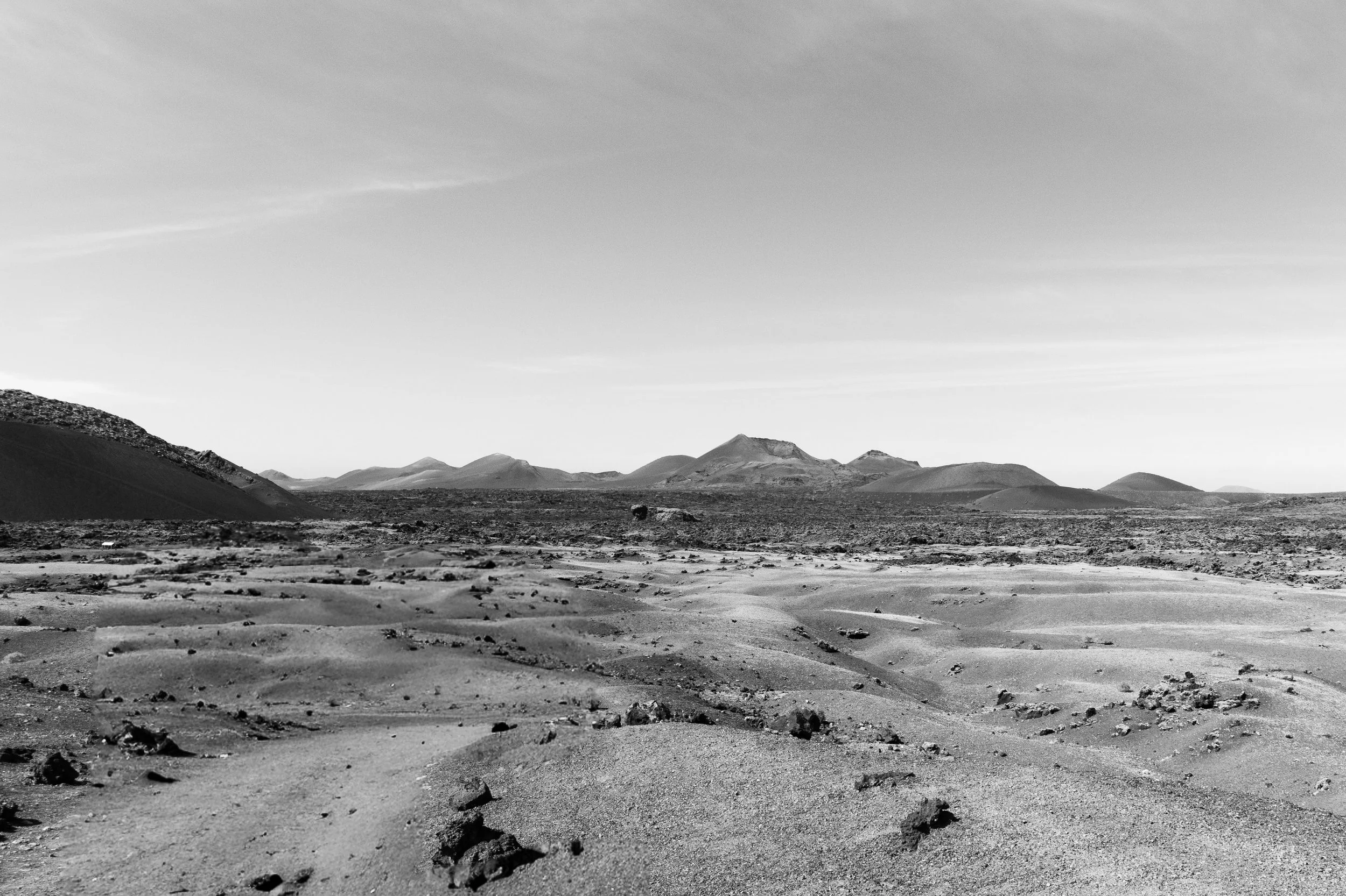 A black and white landscape of a barren desert with sand dunes, rocky terrain, and distant mountains under a clear sky.