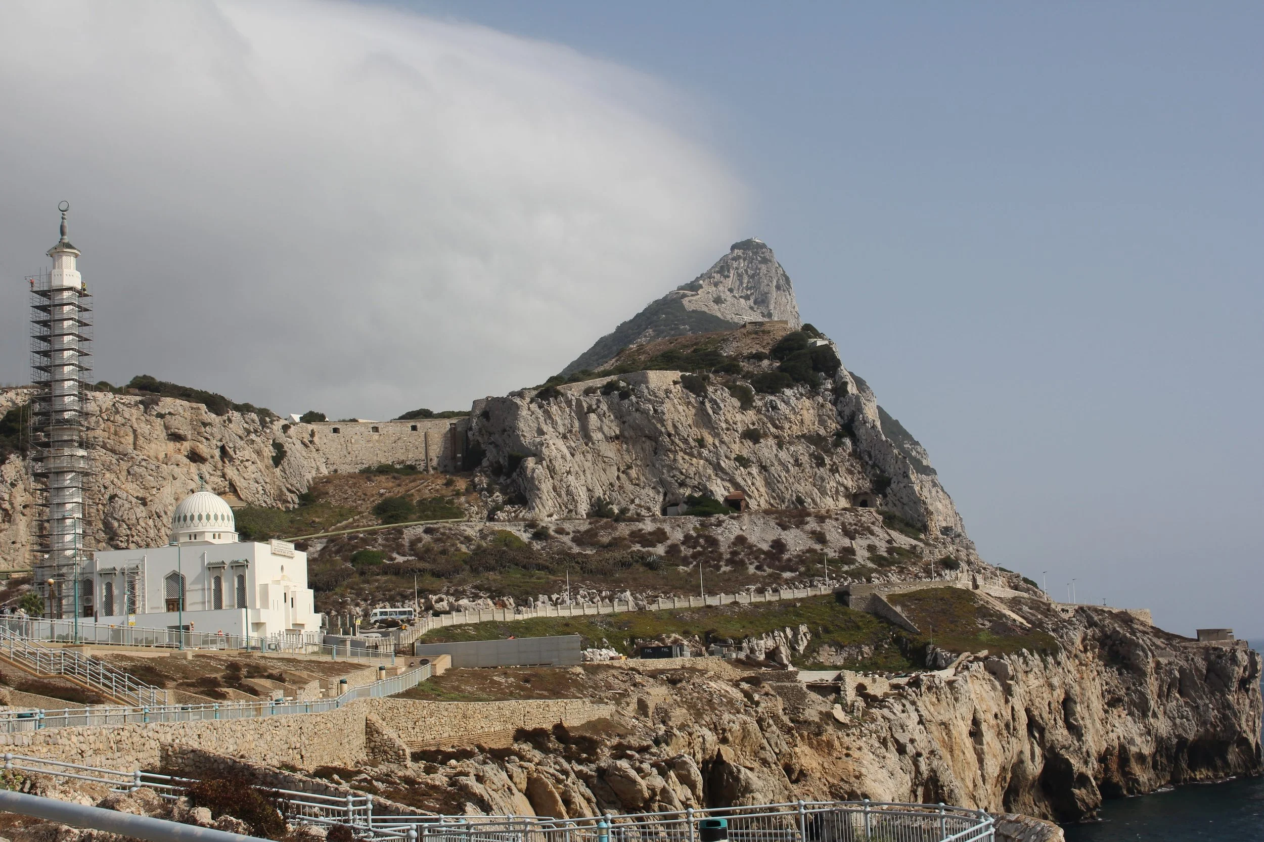Cliffside view of a mountainous landscape with a white chapel, a lighthouse with scaffolding, and winding roads along the rocky terrain.
