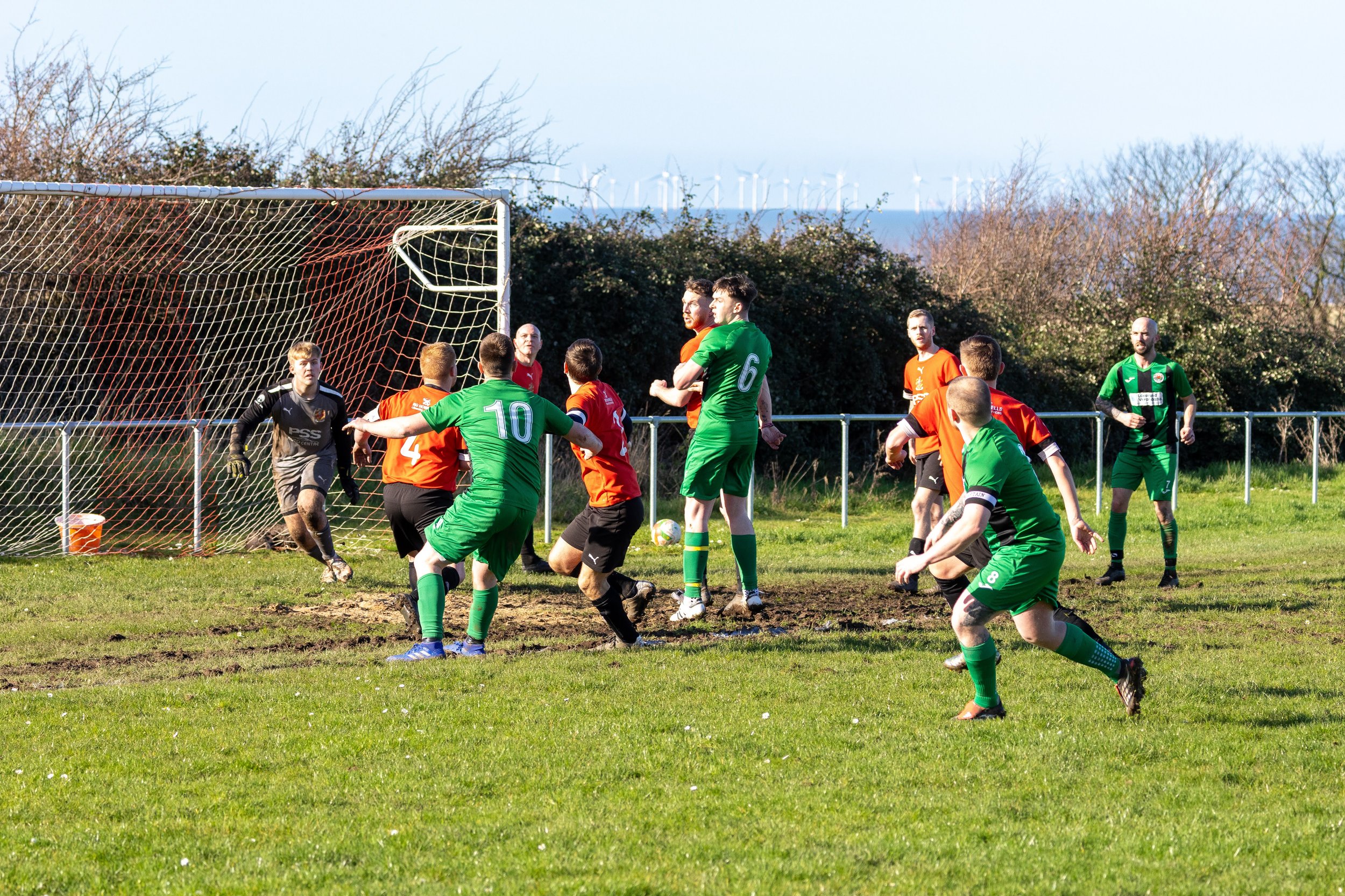 A soccer game in progress with players near the goal, including a goalkeeper in black, players in green and orange jerseys, and a ball near the net. Wind turbines are visible in the background.