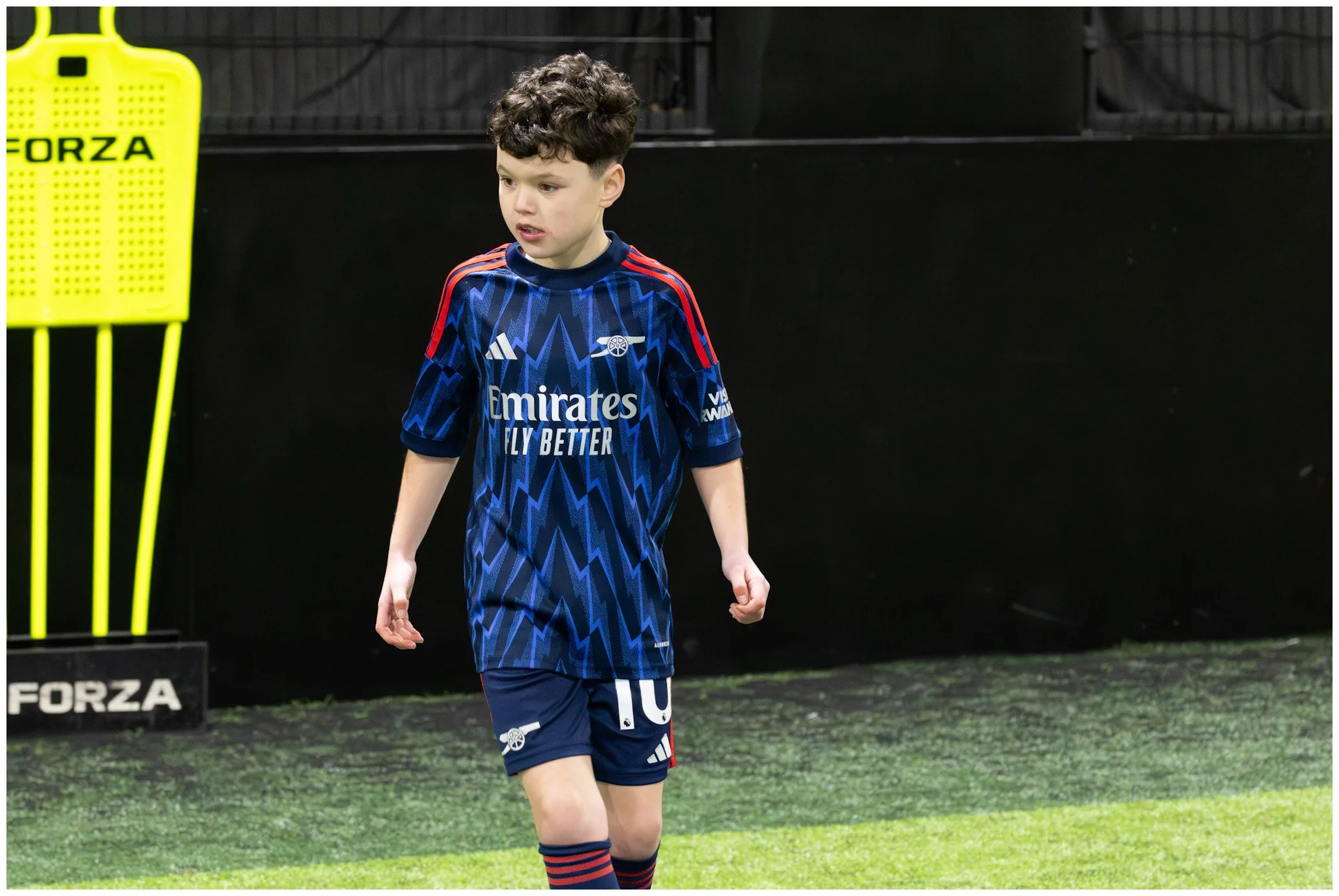 A young boy in a blue soccer uniform with red accents, standing on a soccer field near a yellow training mannequin. The boy appears to be walking or resting during a soccer practice or game.