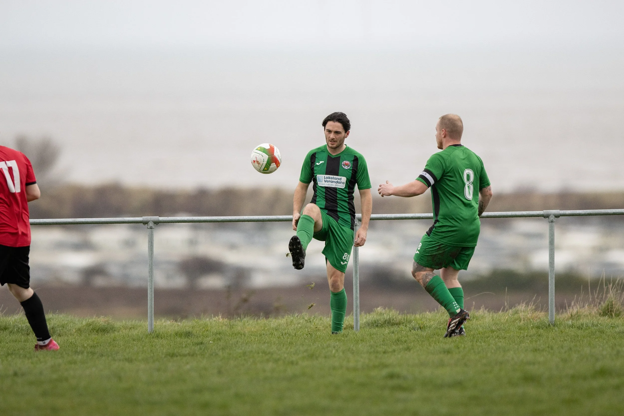 Two soccer players in green uniforms practice near a metal railing on a grassy field, with a gray sky and body of water in the background.