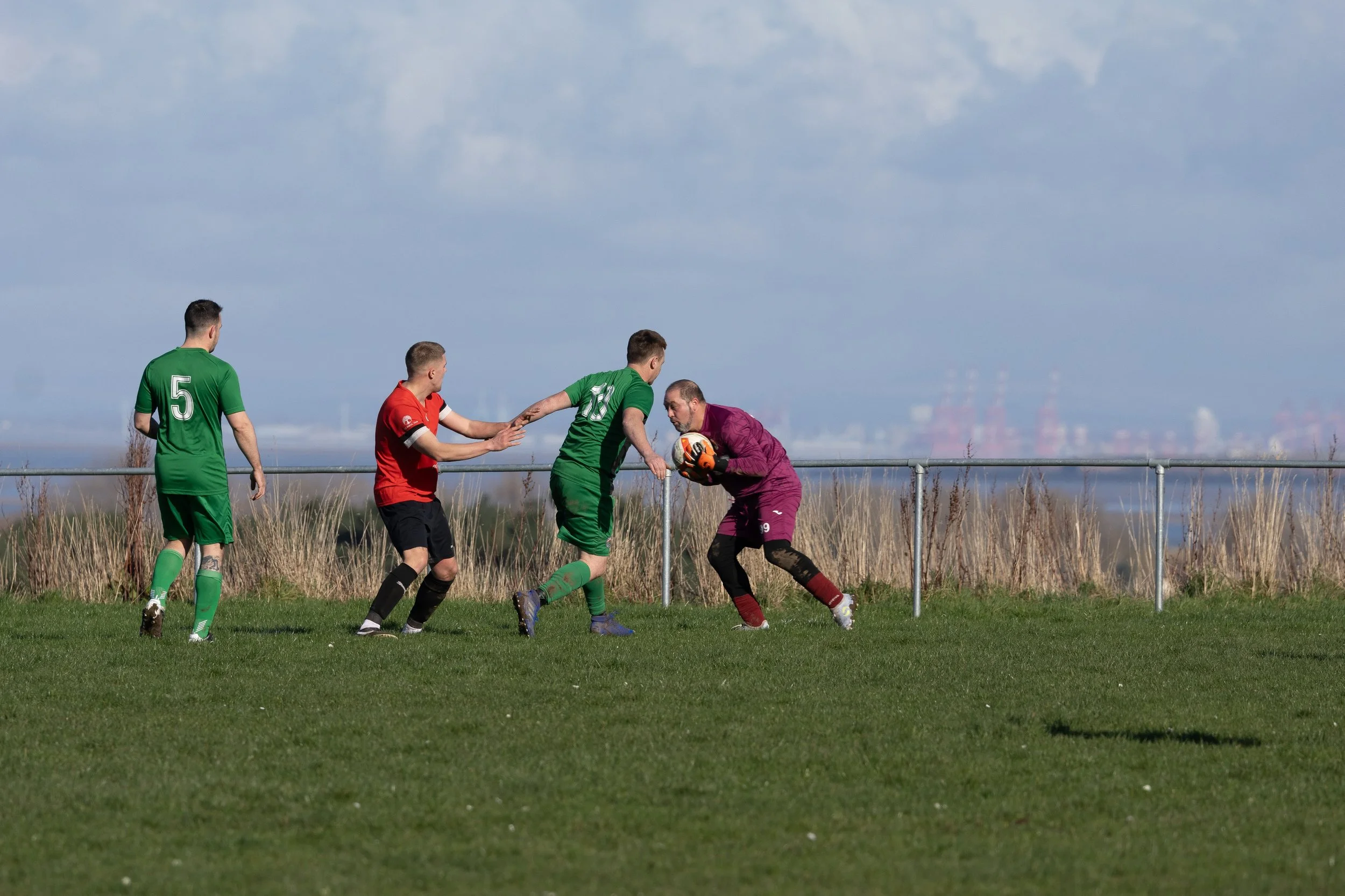 A soccer match with a goalkeeper in purple jersey catching the ball, three players in green and red jerseys approaching, on a grassy field with a fence and field and industrial structures in the background.