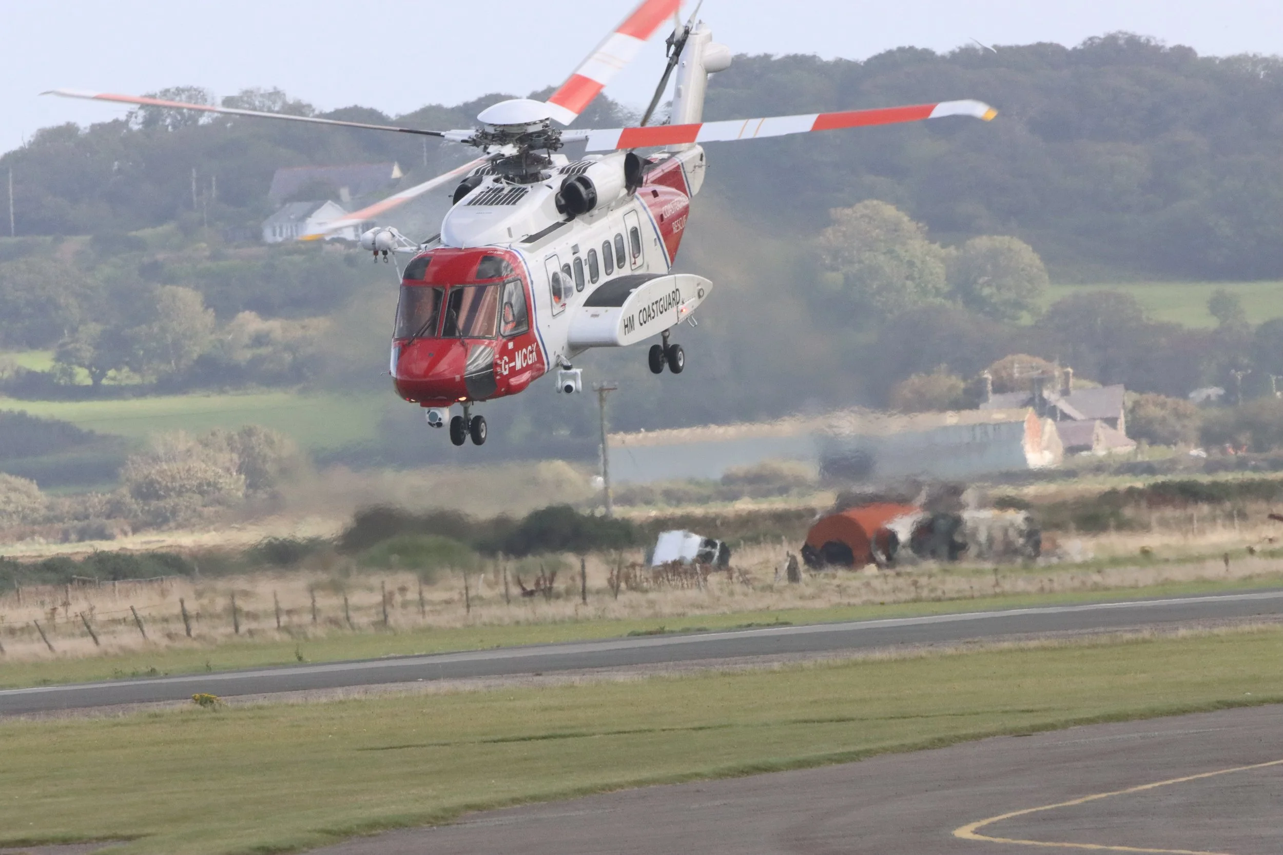 A helicopter in flight near the ground with a damaged vehicle and smoke in the background.