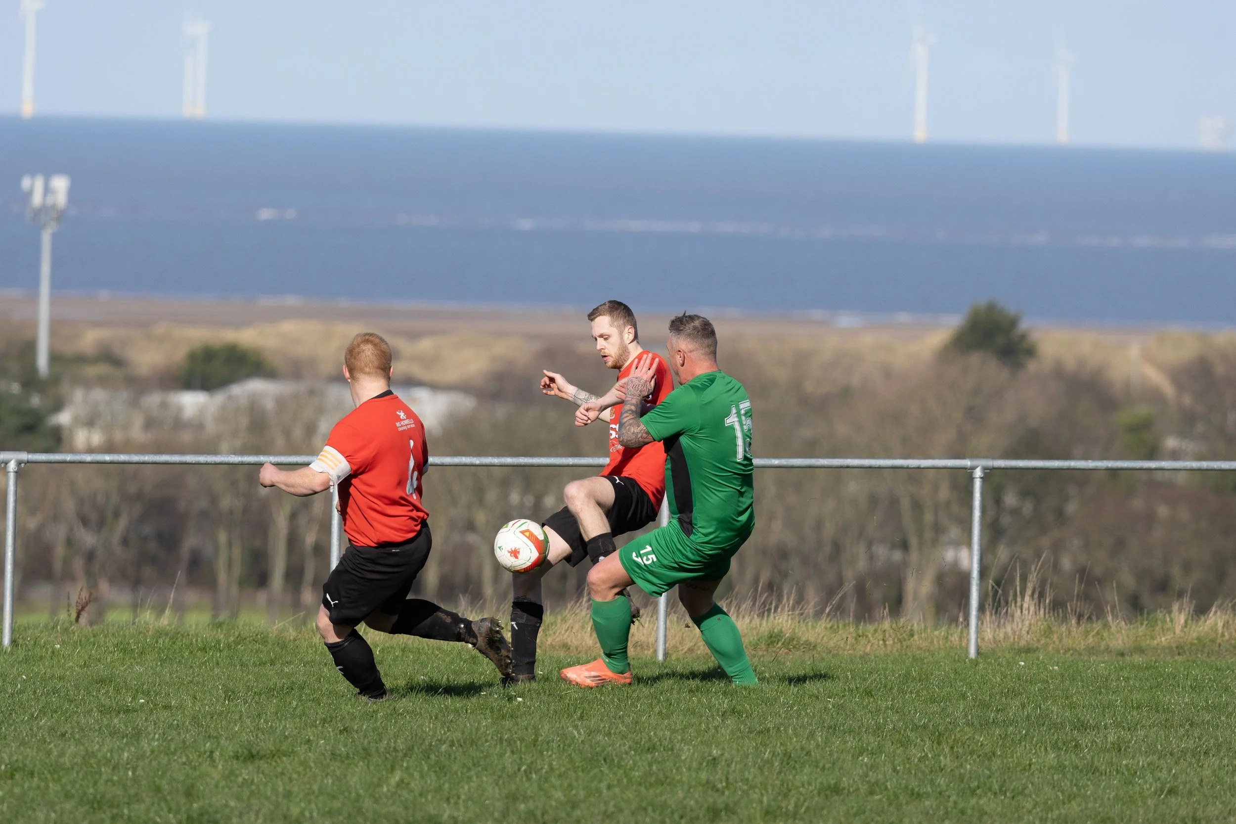 Three soccer players in red and green uniforms competing for the ball on a grassy field with a distant view of water and wind turbines in the background.