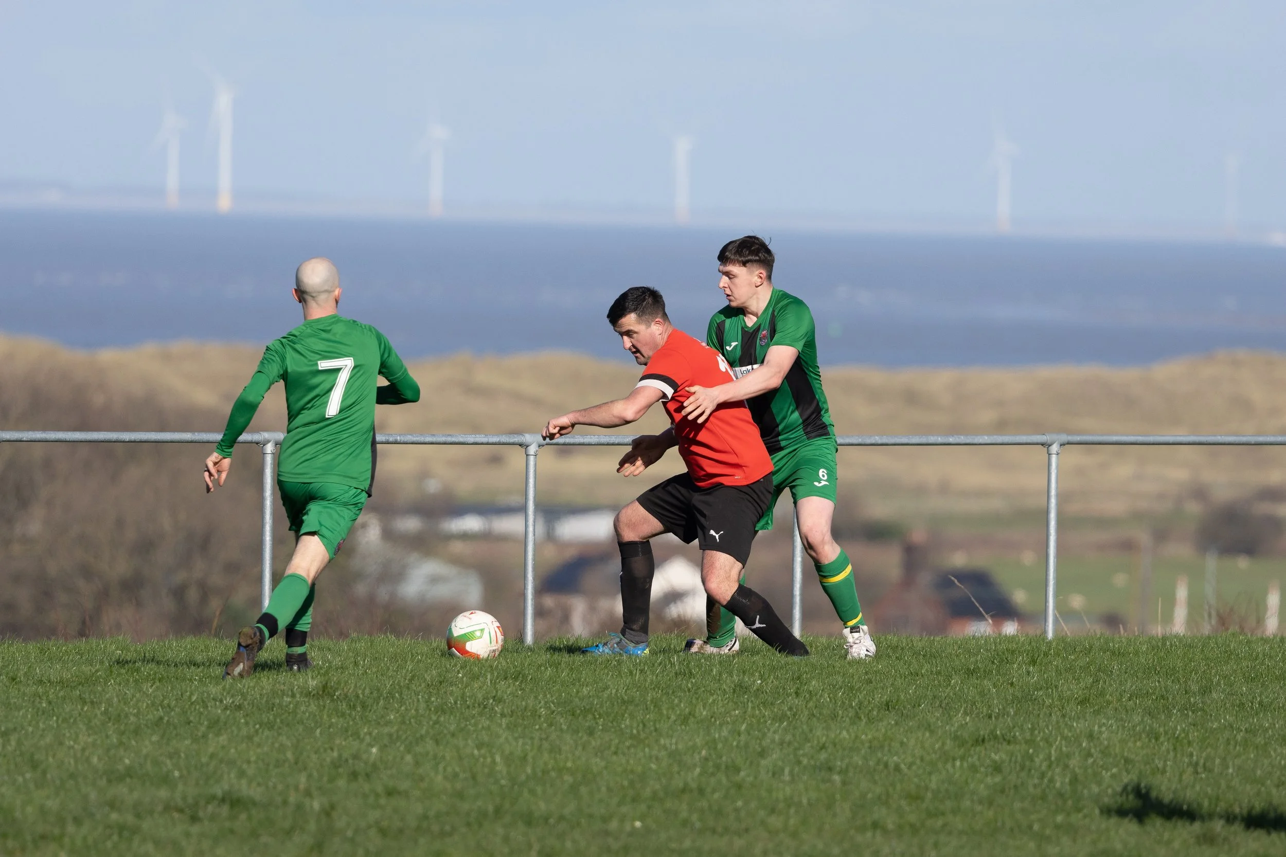 Three soccer players in green and red jerseys competing for the ball on a grassy field with a scenic landscape in the background.