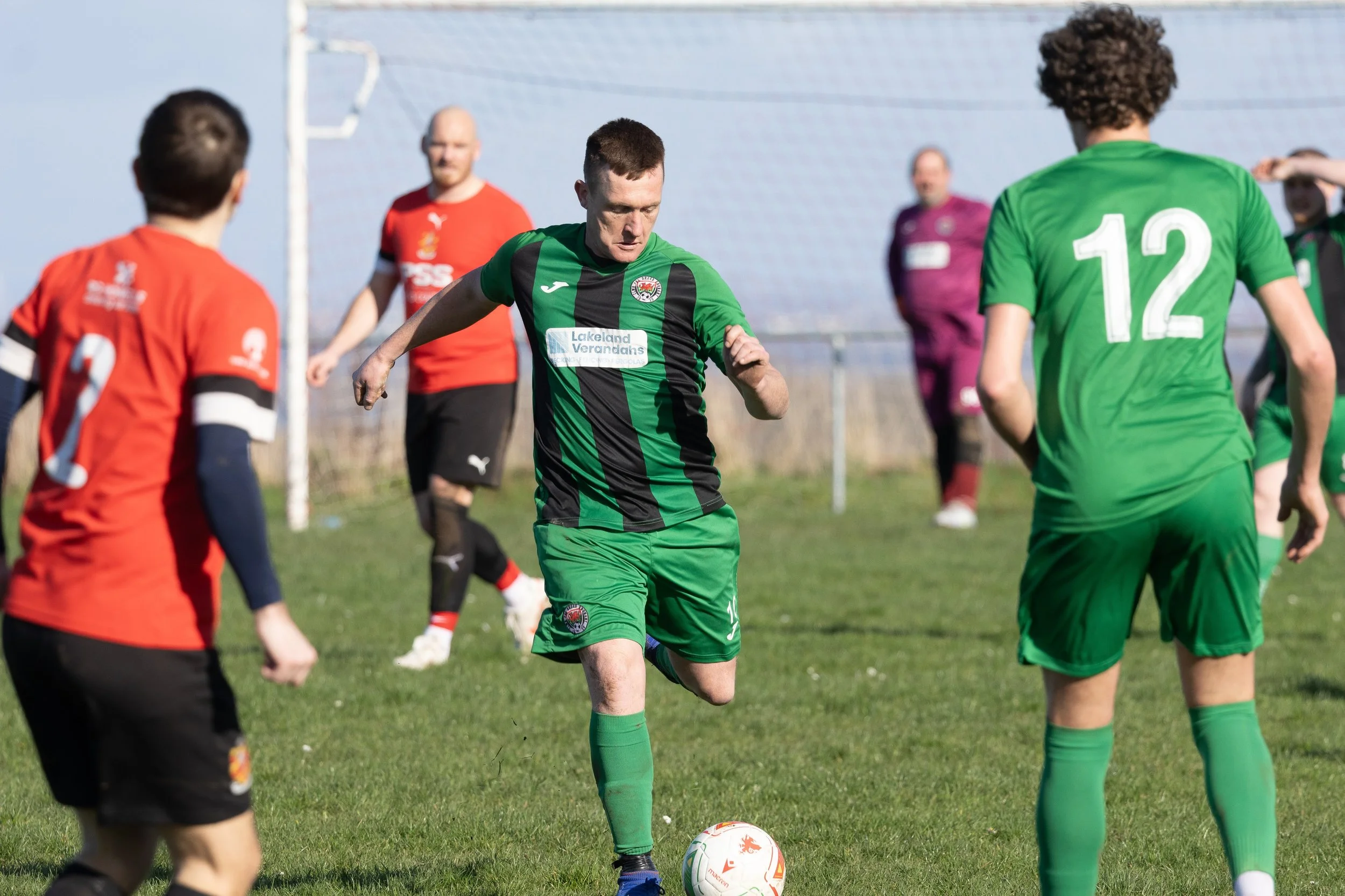Soccer players on a field during a match, with a player in a green and black uniform about to kick the ball, surrounded by players in red and green jerseys and a goalkeeper in a purple kit in the background.