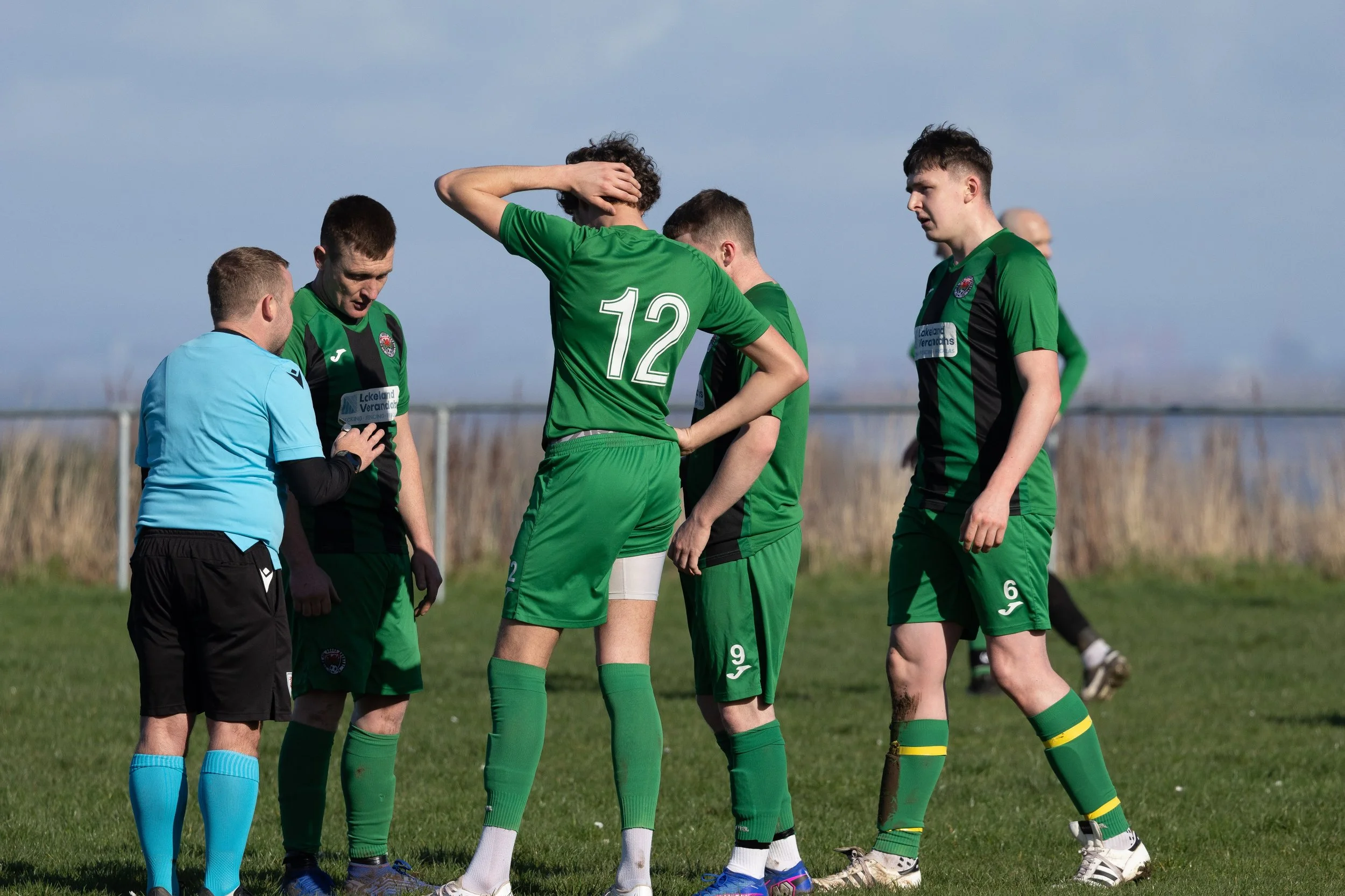 Soccer players in green jerseys huddle on the field, with an assistant referee in blue jersey nearby.