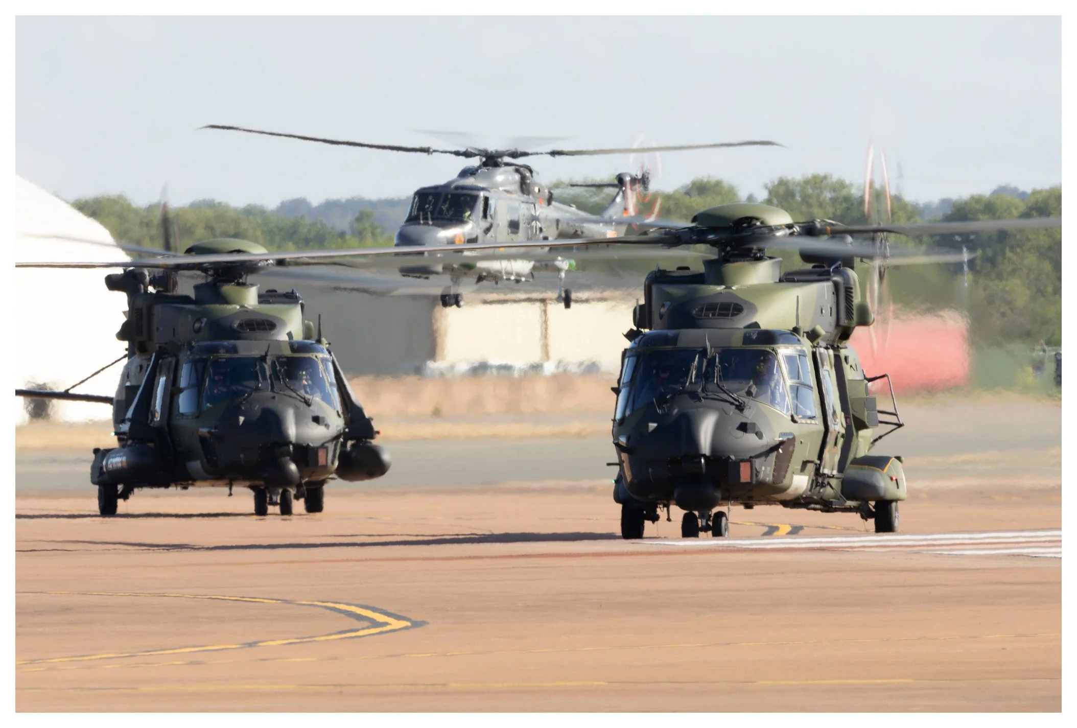 Two military helicopters on the tarmac with a military helicopter in the background.