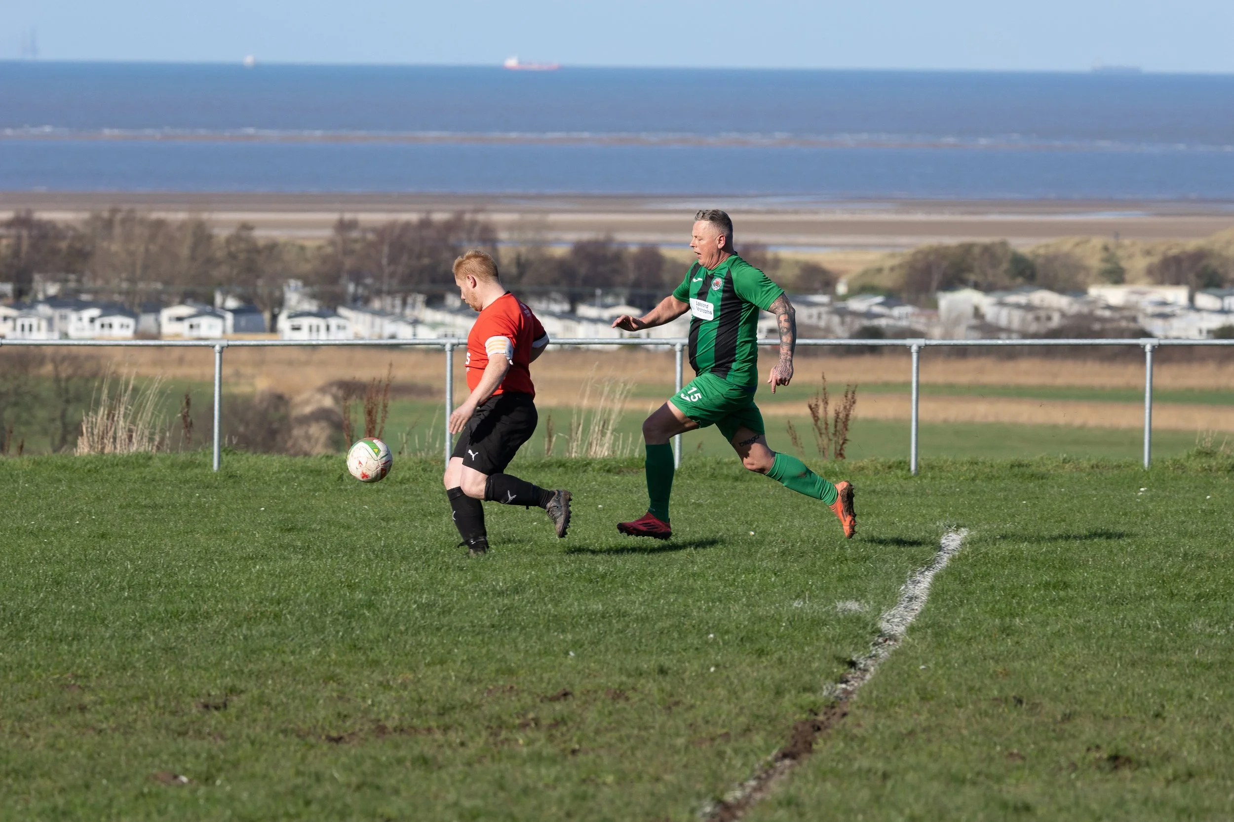 Two soccer players on a grassy field, one in a red and black uniform and the other in a green uniform, competing for the ball during a game, with a body of water and a landscape in the background.