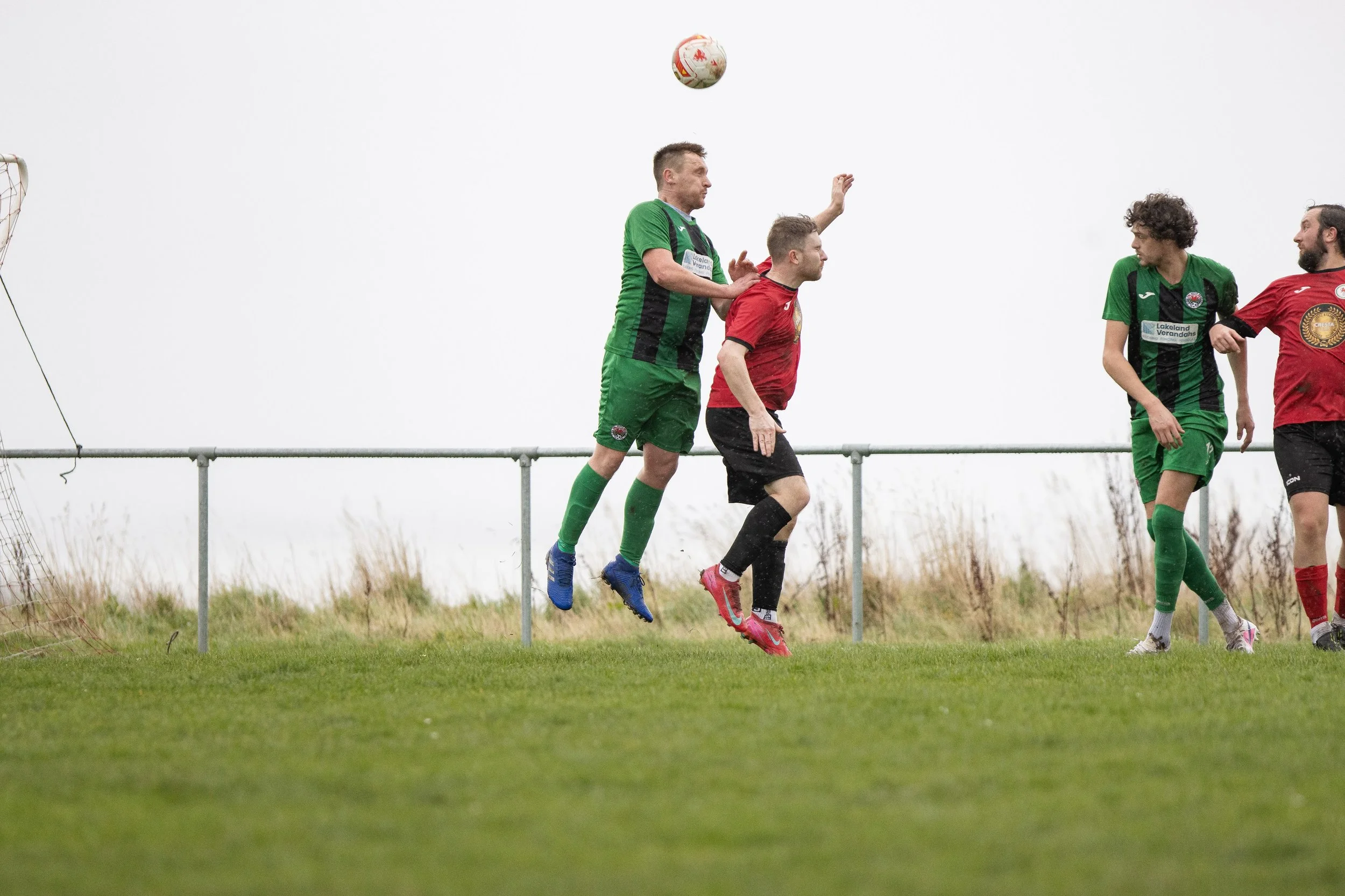 Four soccer players, two in green uniforms and two in red uniforms, competing for a header over a grassy field.
