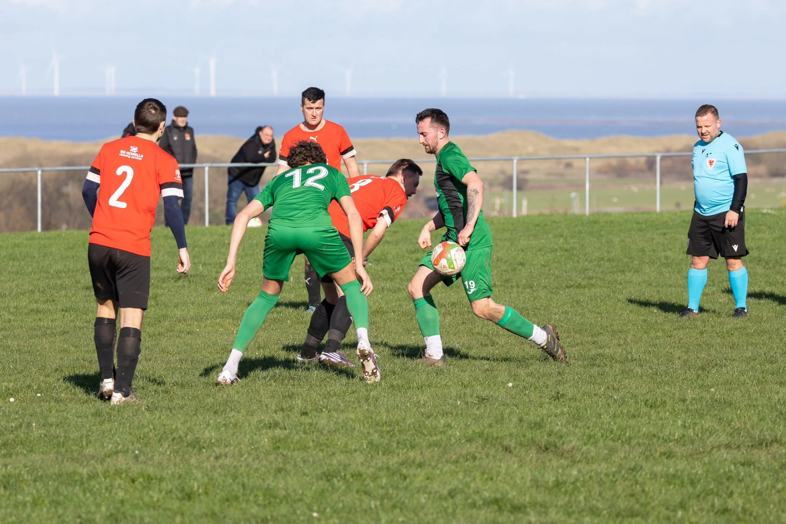 Soccer match where players in orange and green jerseys are competing for the ball on a grassy field, with the referee in a blue jersey observing.