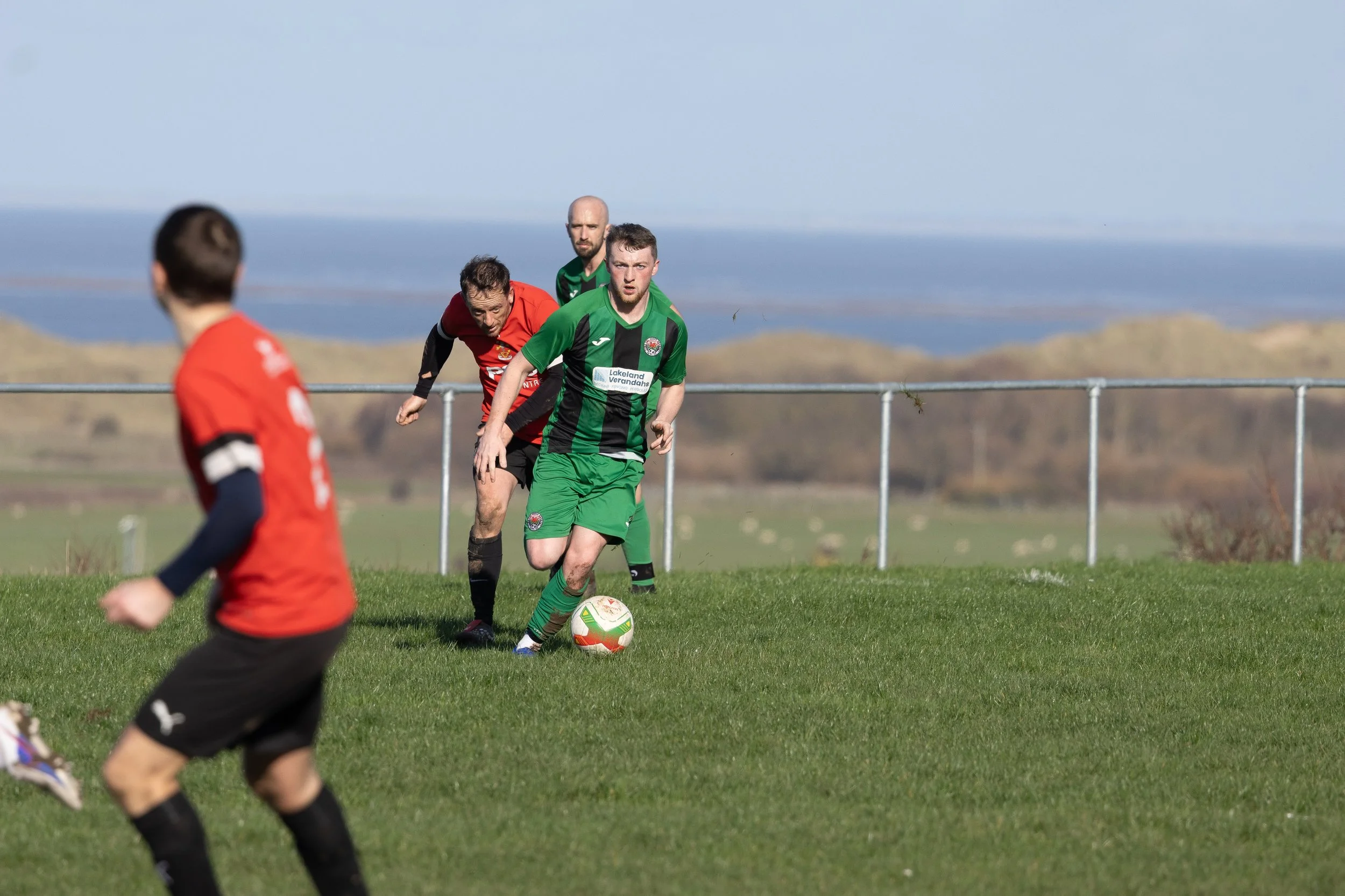 Soccer players in green and red uniforms playing on a grassy field with a scenic backdrop of hills and water.