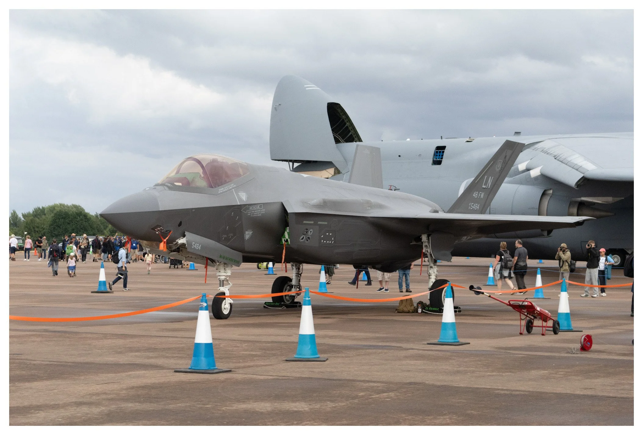 A gray military fighter jet on display at an air show, surrounded by orange and blue traffic cones, with spectators walking around in the background. A large aircraft is visible behind the jet.