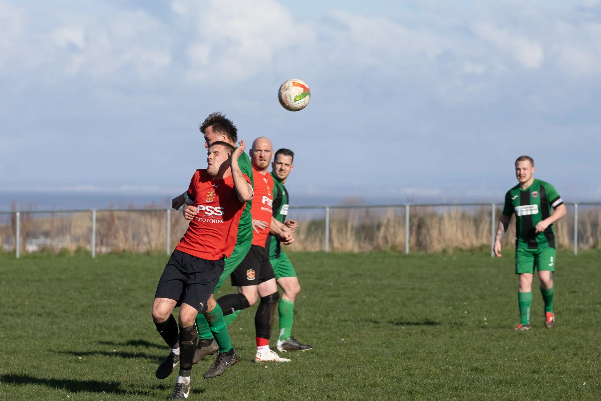 Soccer players competing for the ball on a grassy field, with four players visible, dressed in red and green uniforms, under a partly cloudy sky.