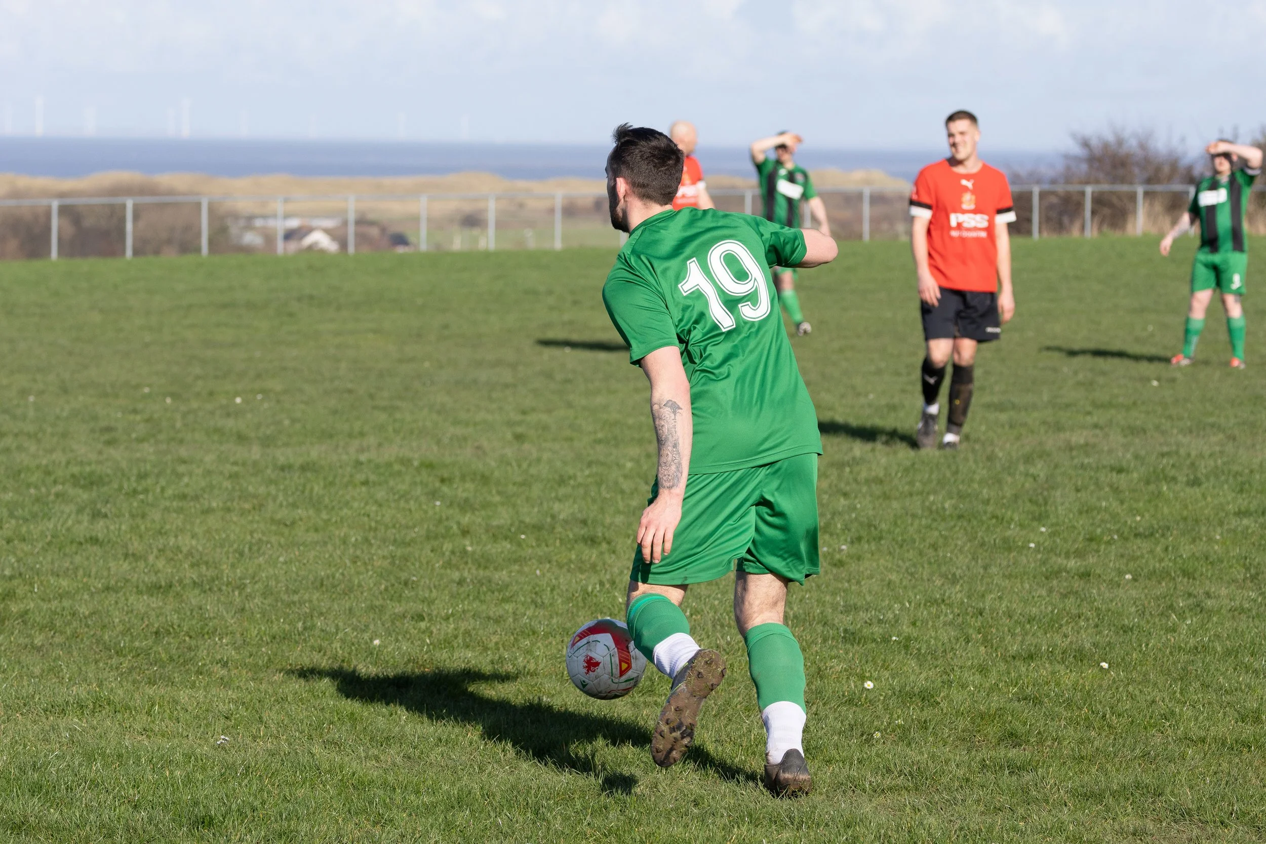 A soccer player in a green uniform with the number 19 is about to kick a soccer ball on a grassy field. In the background, other players and a fence are visible, with a blue sky and distant landscape.