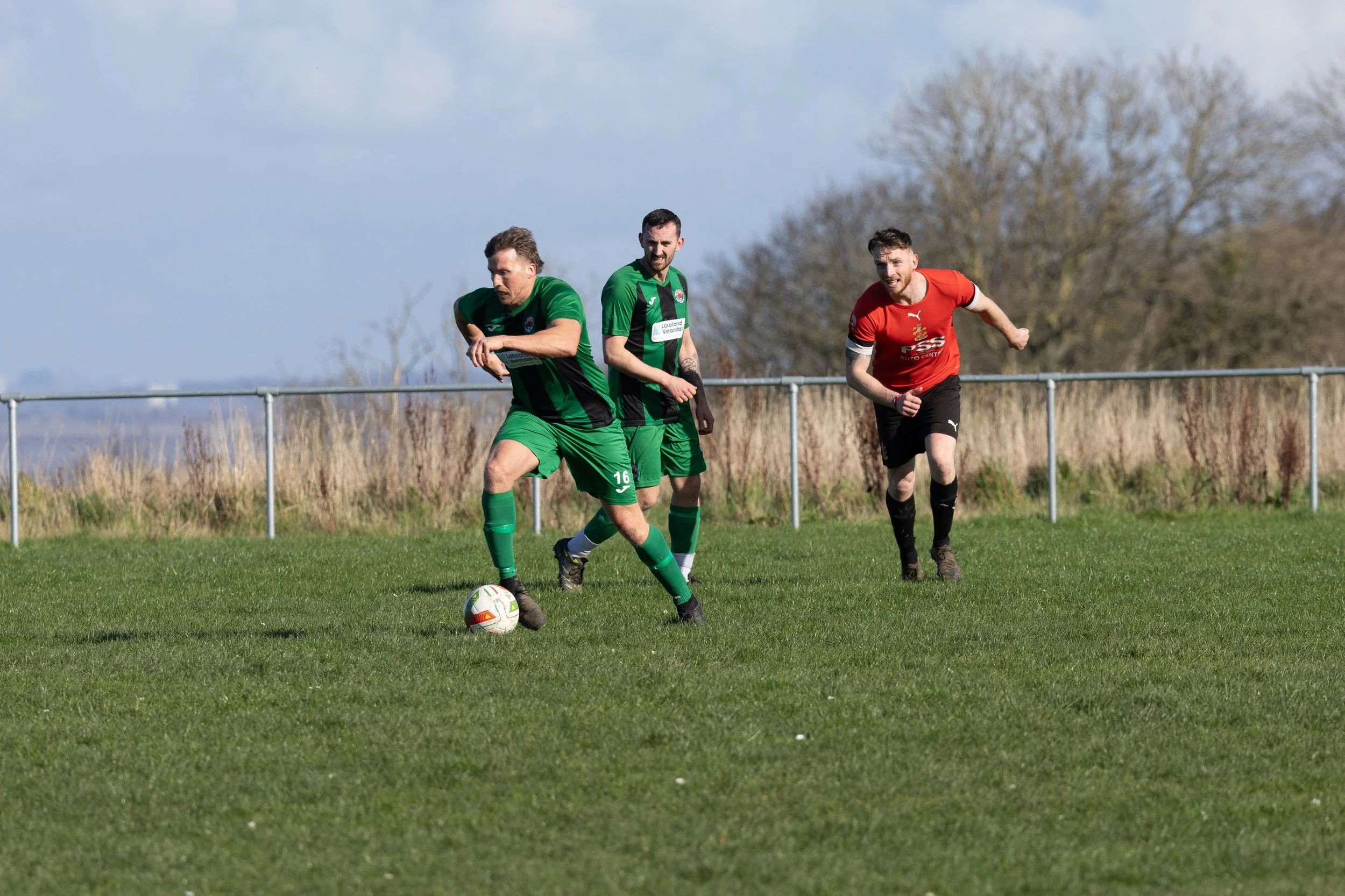 Three soccer players playing on a grassy field, two in green jerseys and one in red, with a background of trees and a cloudy sky.