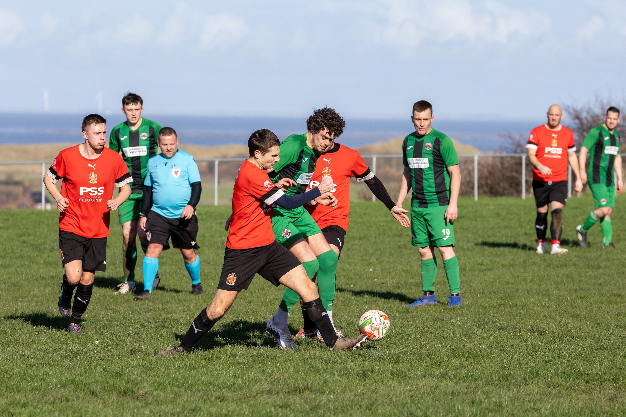 Soccer match with players in green and red jerseys competing for the ball on a grassy field under a cloudy sky.