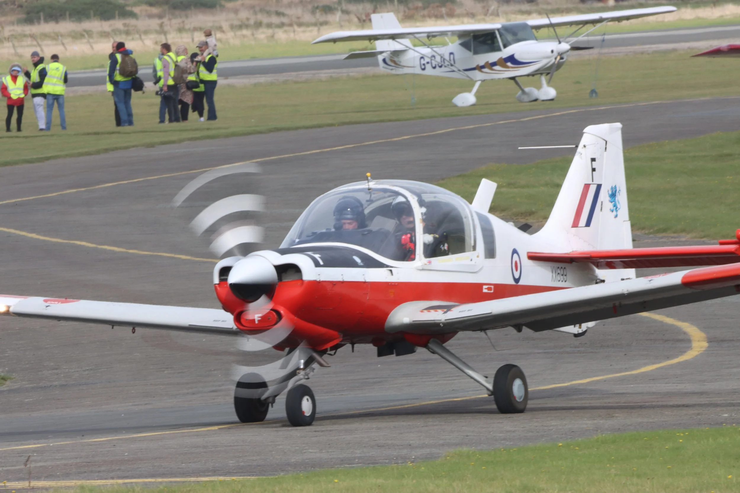Small red and white jet aircraft taxiing on runway, with crew members inside cockpit. In background, people in yellow safety vests and casual clothing observing, and a light aircraft parked behind a grassy field.