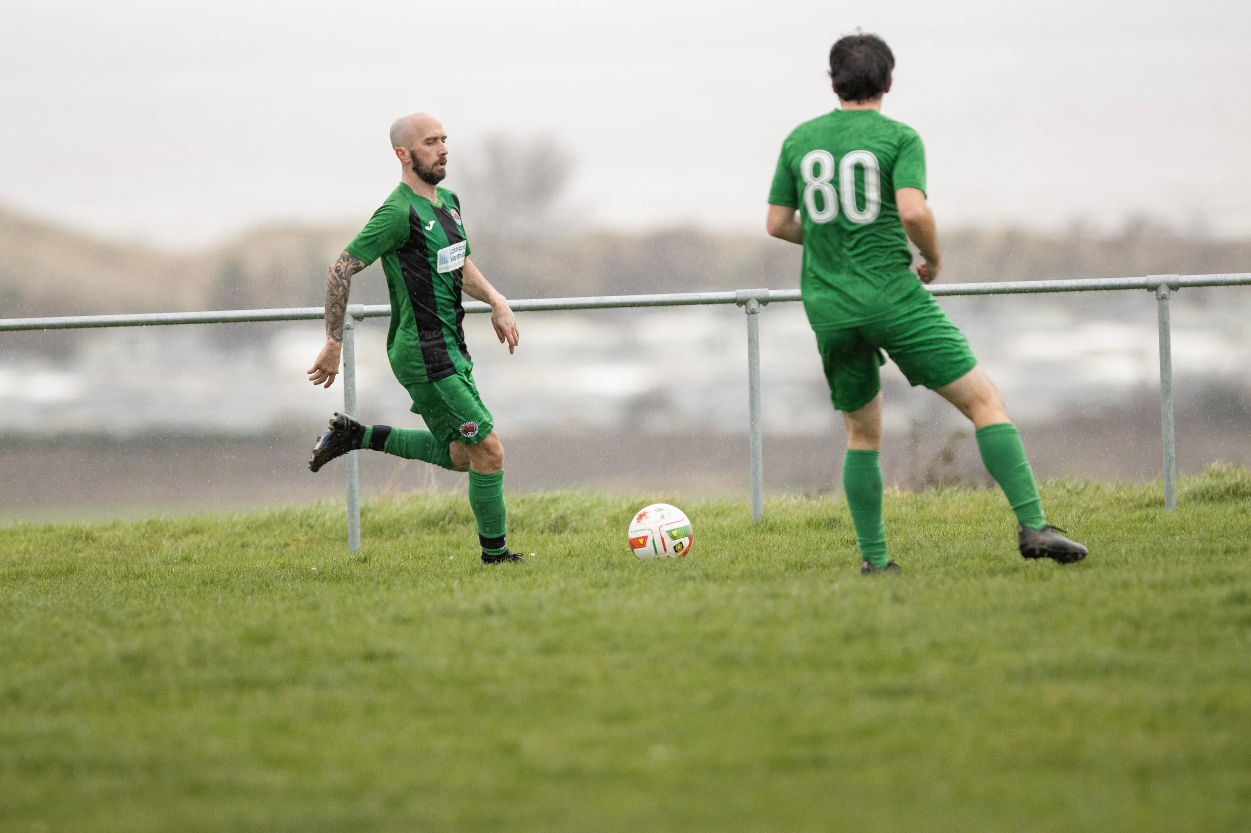 Two soccer players in green uniforms practicing on a grassy field near a metal railing, with an overcast sky and blurred background.