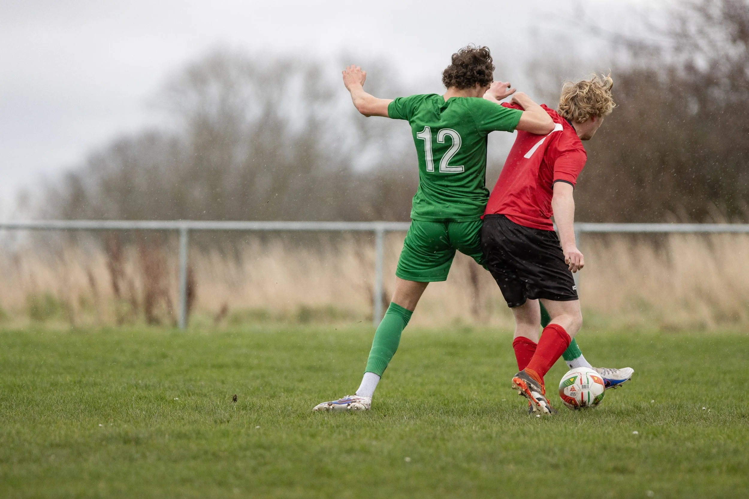 Two young soccer players competing for the ball during a match on a grass field in rainy weather.