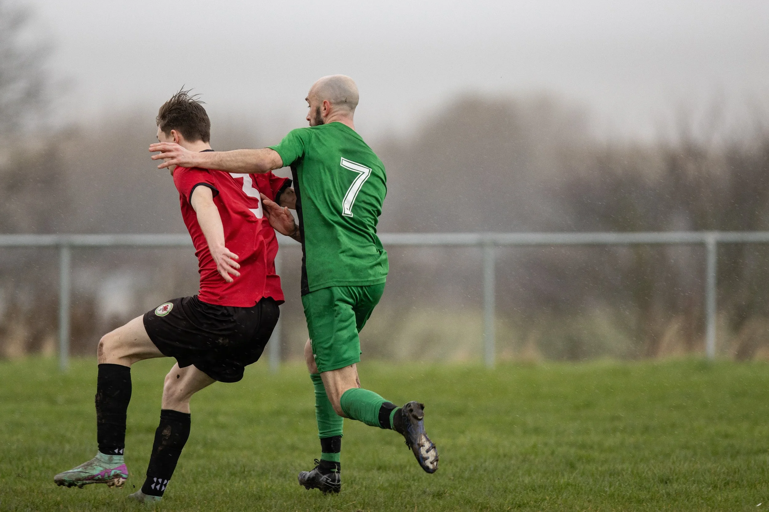 Two soccer players, one in a red jersey and the other in a green jersey with the number 7, are competing for the ball on a grassy field. The player in red is attempting to kick the ball while the player in green is trying to block or tackle. The back