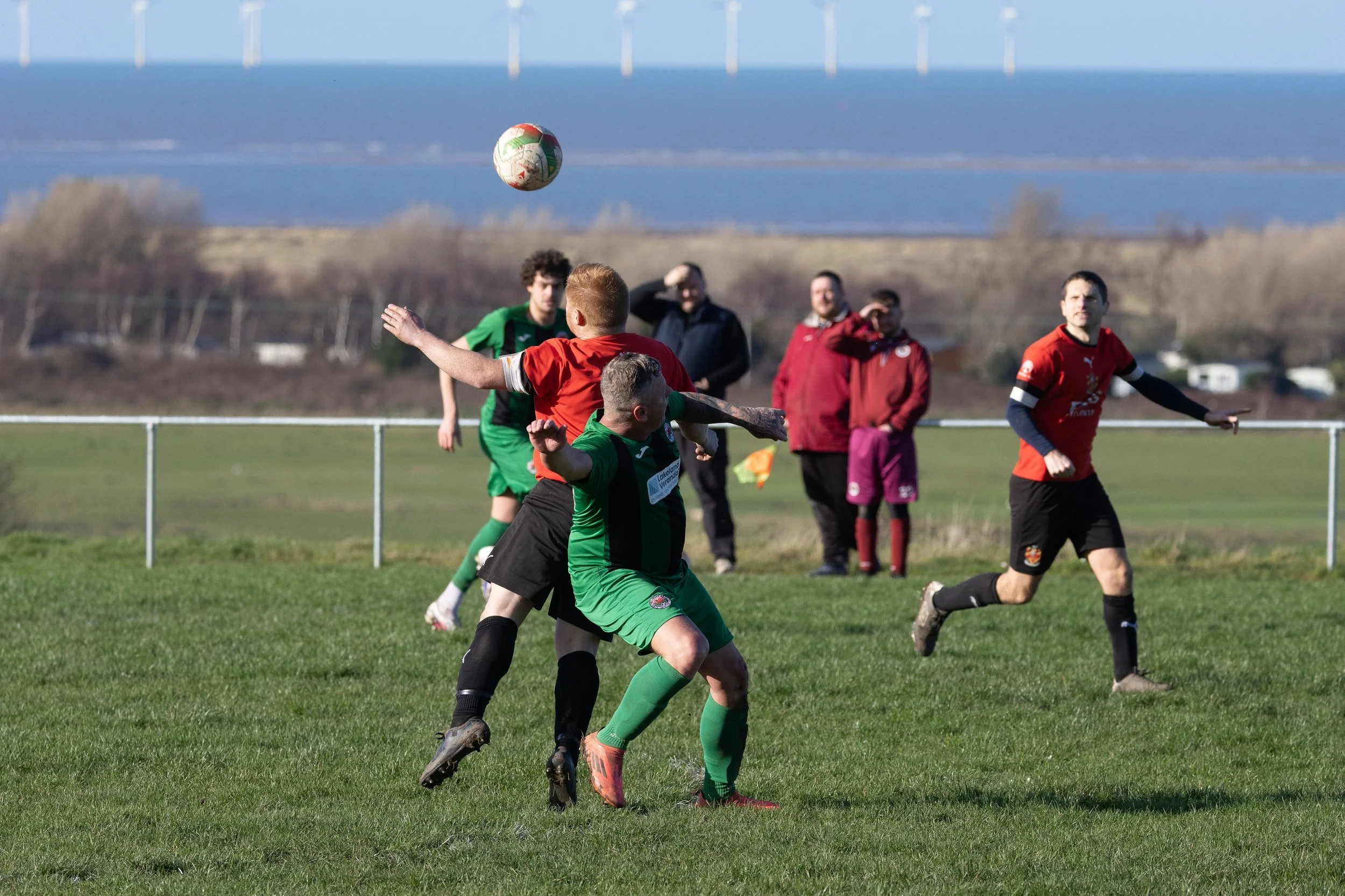 Soccer players in green and red uniforms contesting for the ball during a match on a grassy field with a scenic background of hills and sky.