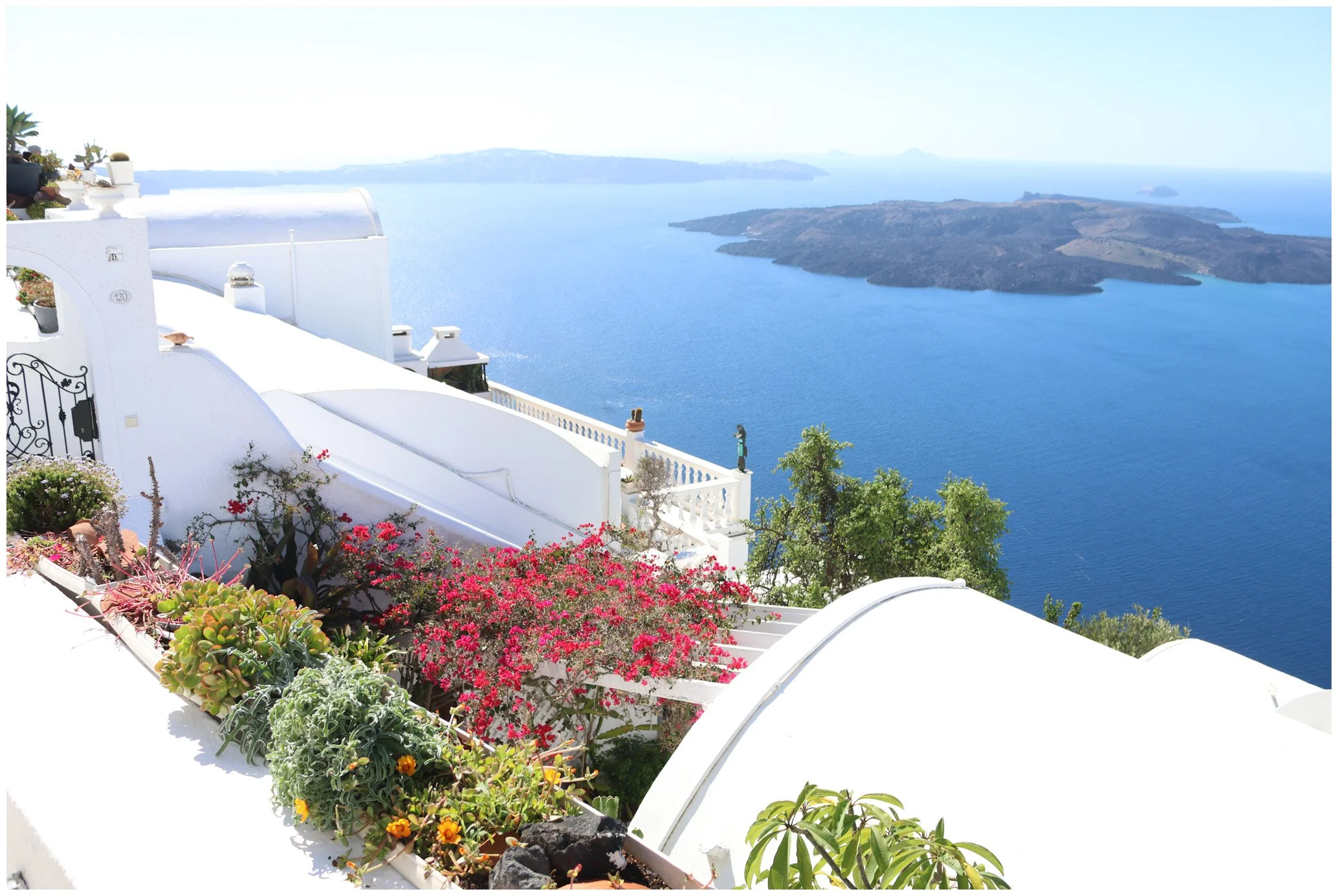 White buildings with curved roofs and terraces overlooking a deep blue sea and distant islands, with colorful potted plants and flowers in the foreground.