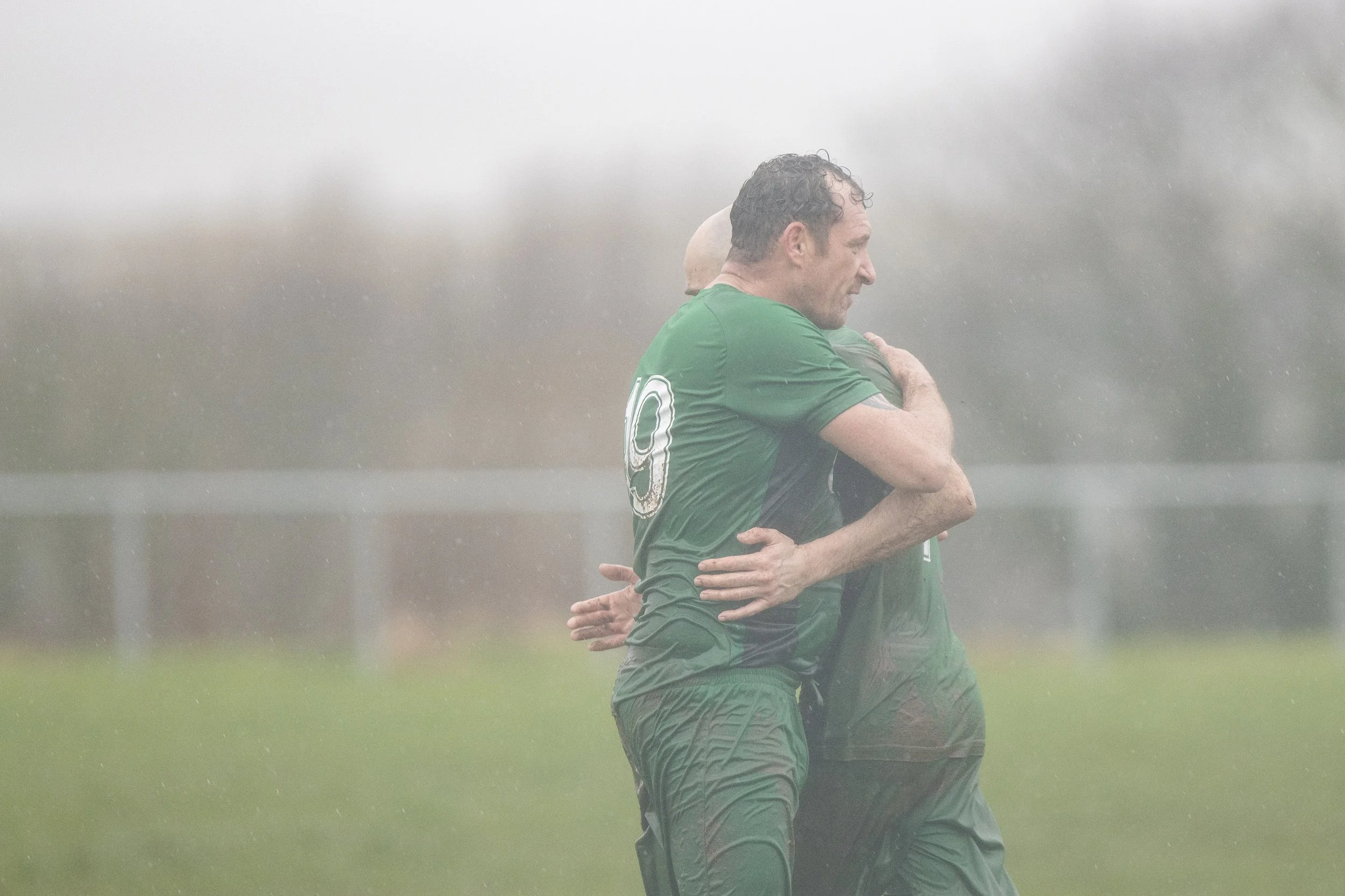 Two soccer players embracing in the rain on a field
