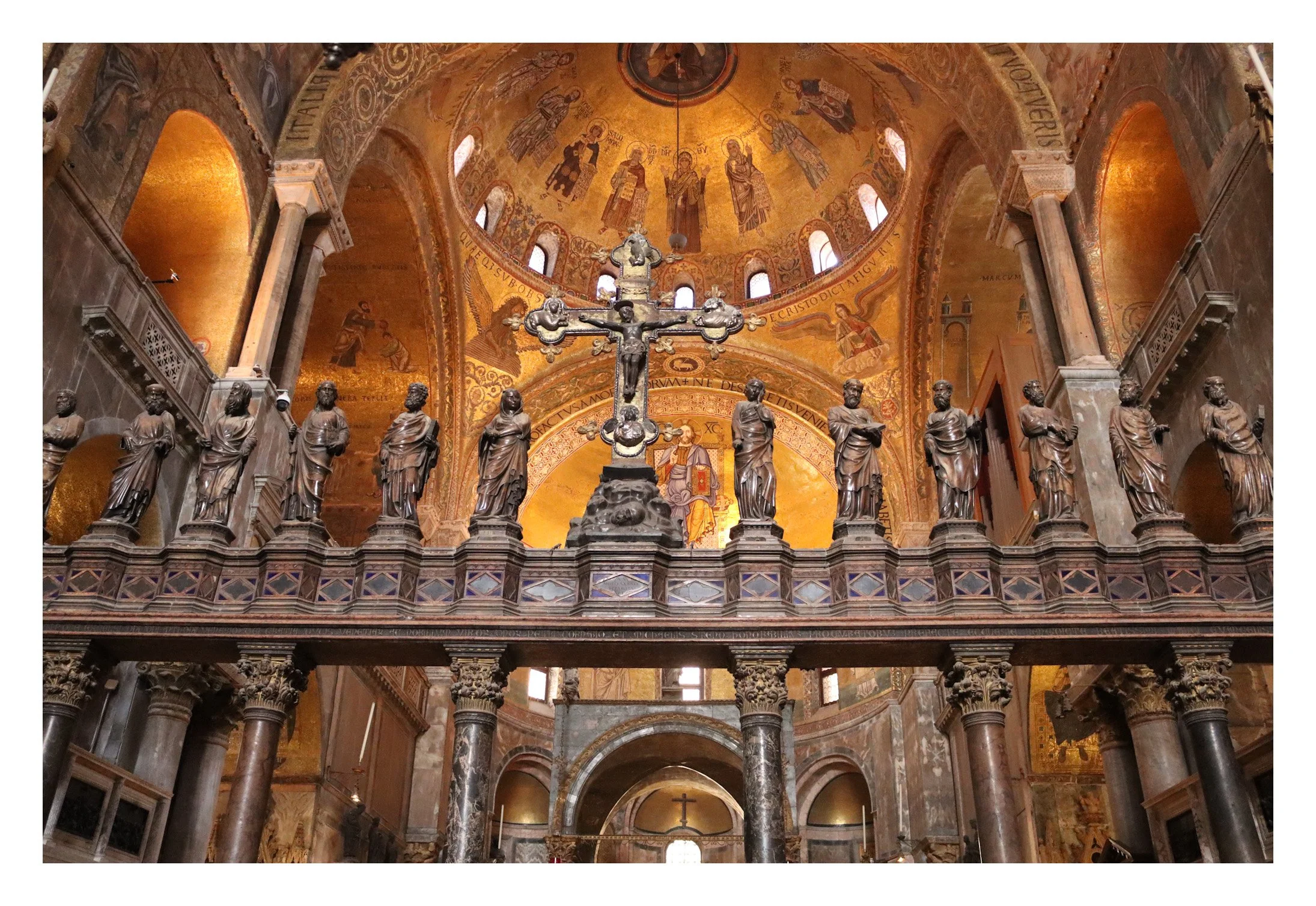 Inside a historic church with a gold mosaic ceiling, a large crucifix with Jesus on the cross, and a row of bronze statues of saints or apostles on a balustrade.