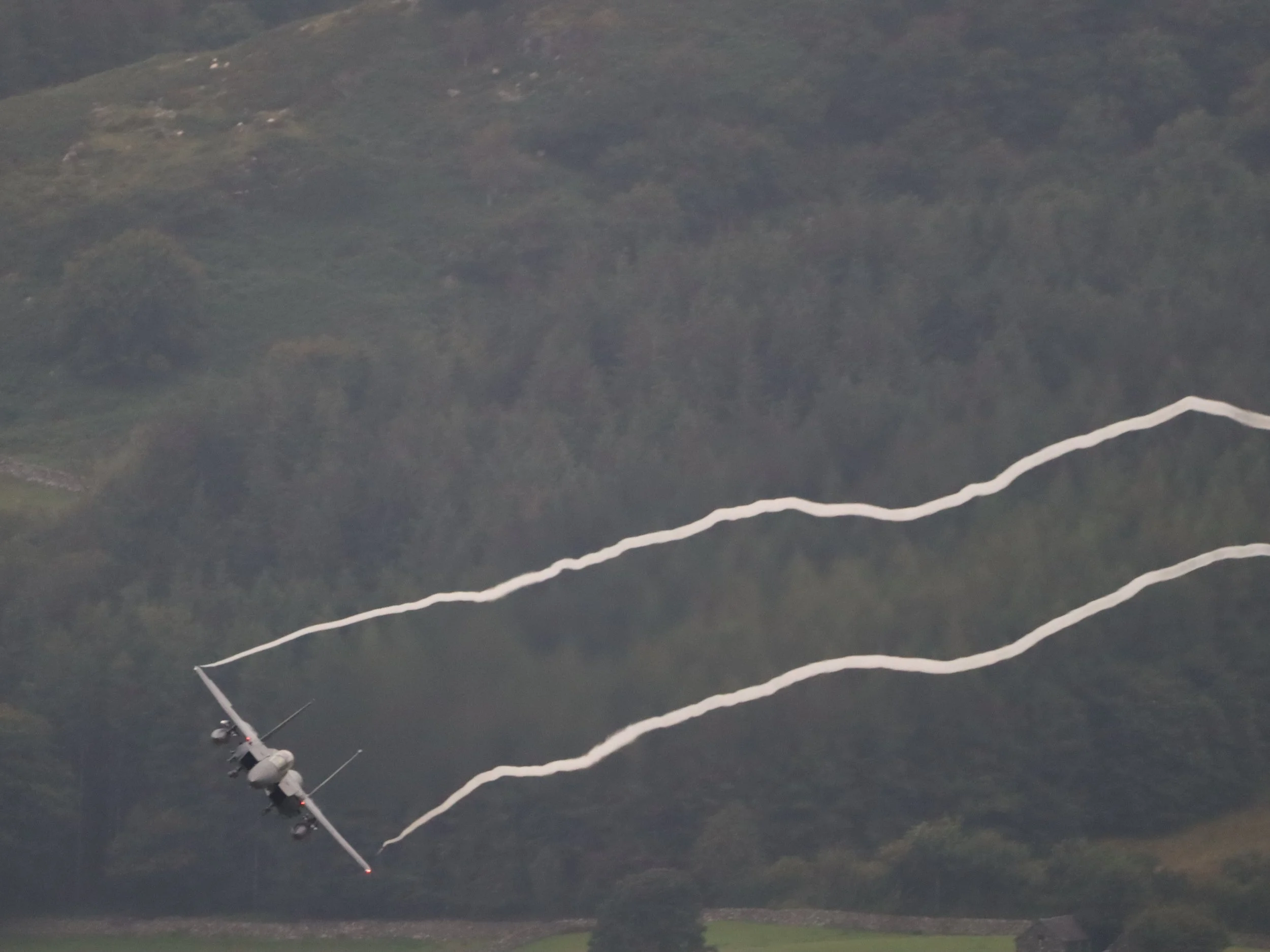 A military aircraft flying over a dense forest with visible white contrails behind it.