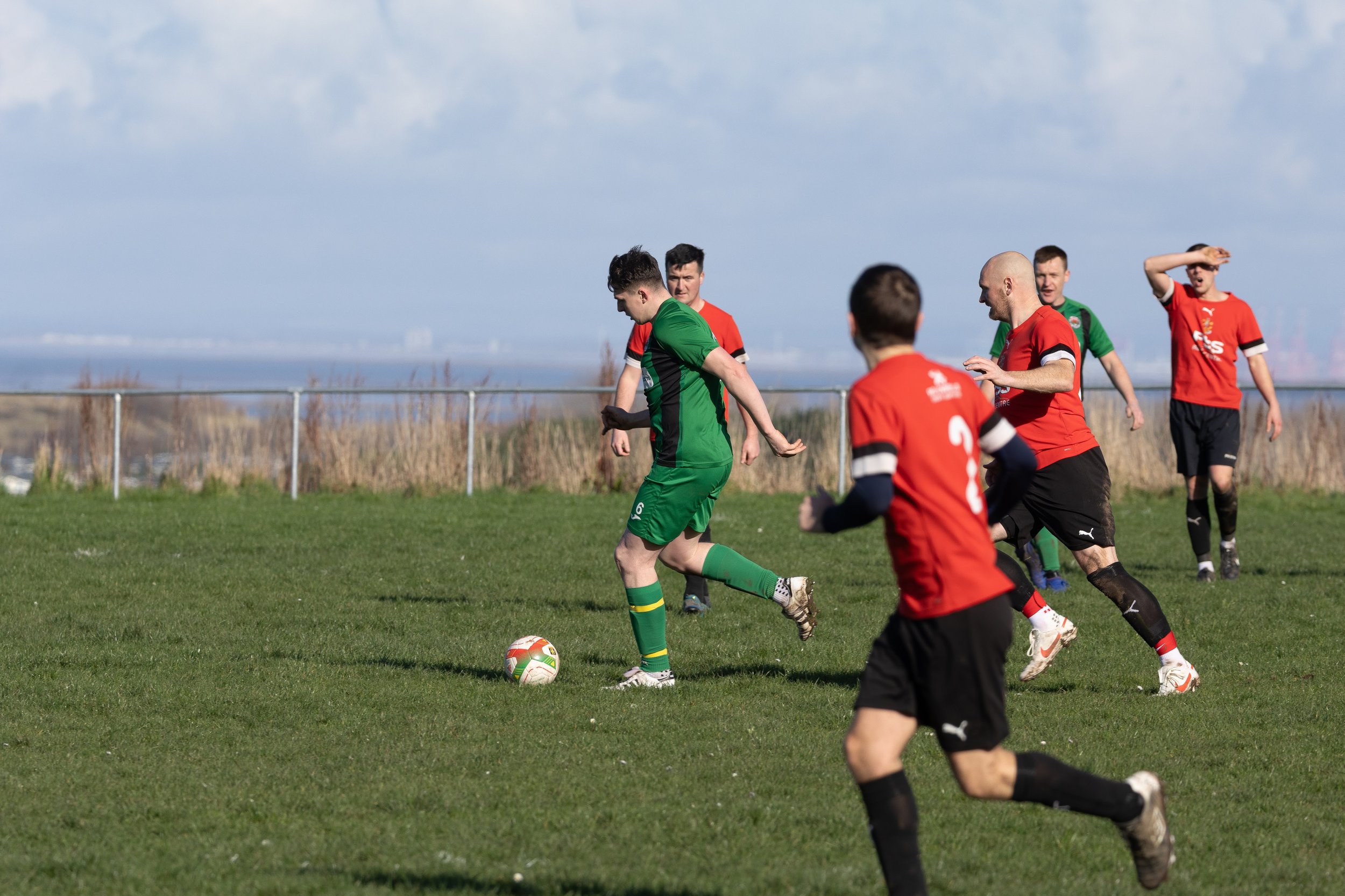 Soccer match with players in red and green jerseys on a grass field, with a clear sky and open landscape in the background.