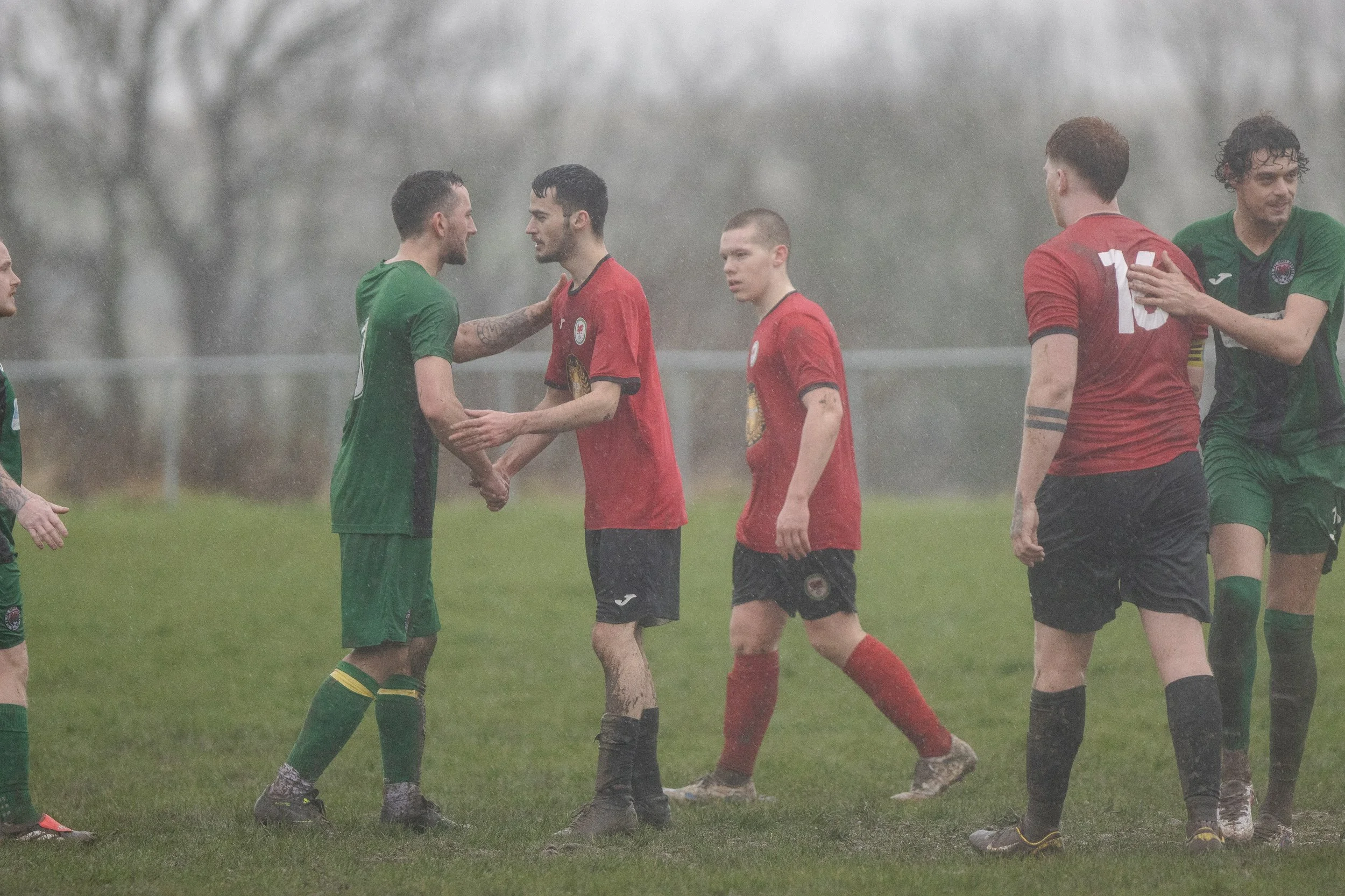 Soccer players shaking hands on a rainy field, some with wet hair and muddy clothing, in front of foggy trees.