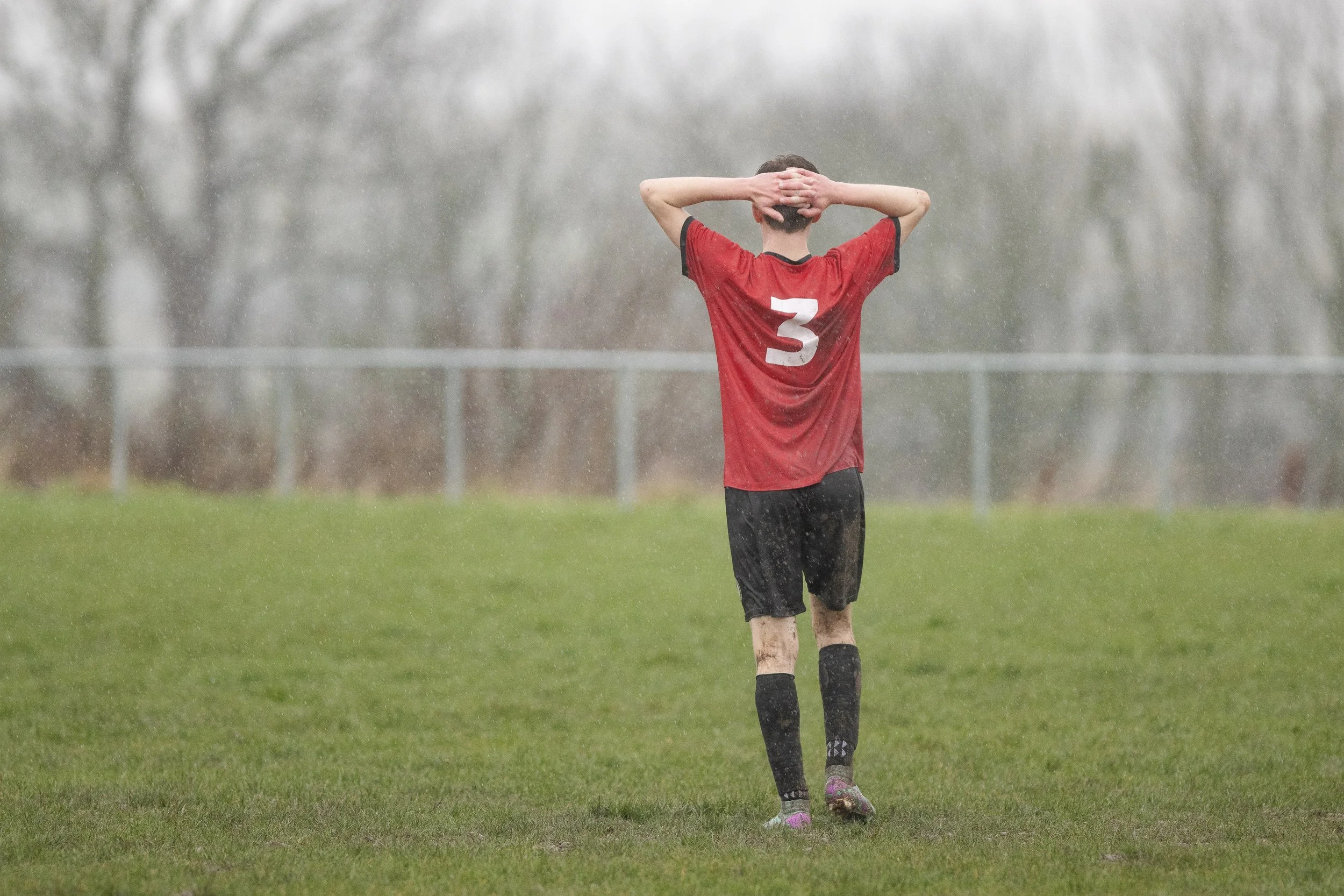 Soccer player in a red jersey with the number 3, standing on a wet green field with his hands on his head, during rainy weather.