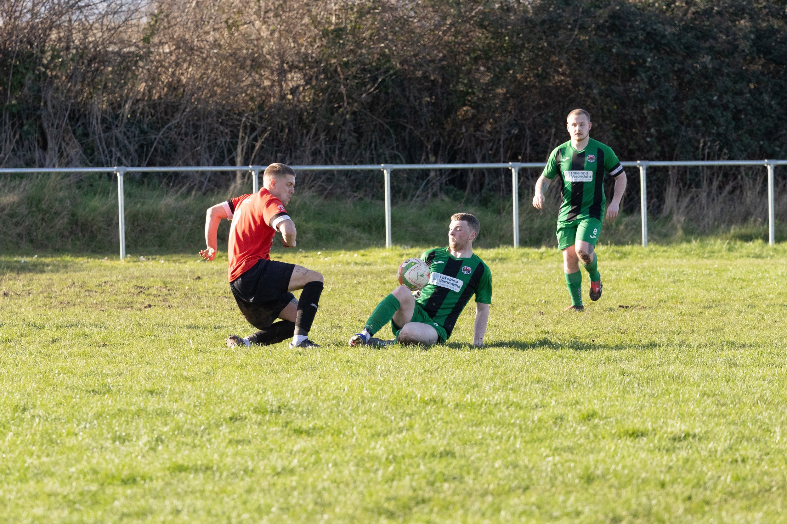 Three male soccer players on a grassy field during a match, with one player in a red jersey and two players in green and black jerseys. Two players are involved in a play with a white soccer ball, one sitting on the ground and the other kneeling. The