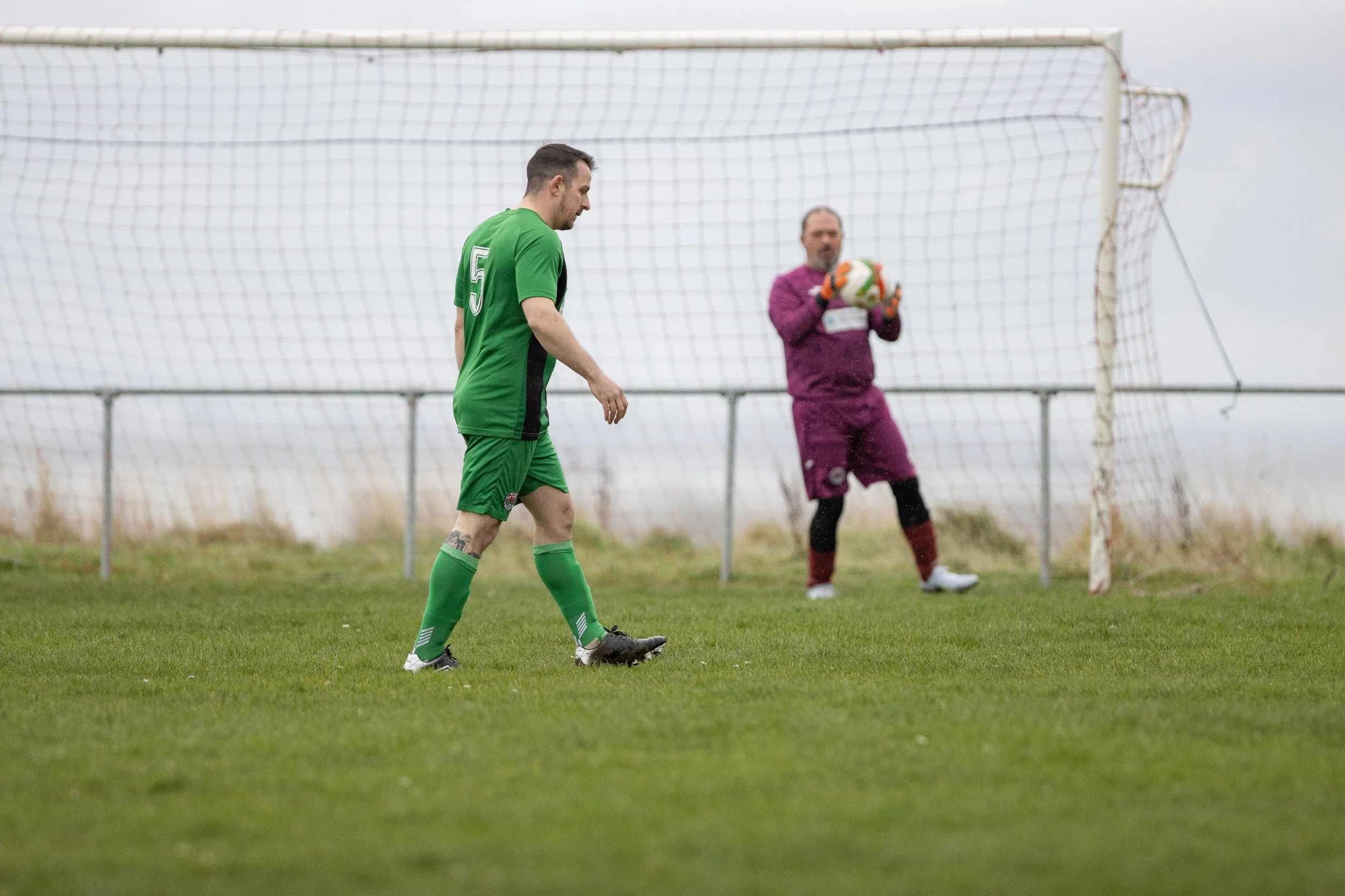 A soccer player in a green uniform walking on the field with a goalkeeper in a purple uniform holding a soccer ball near the goal net.