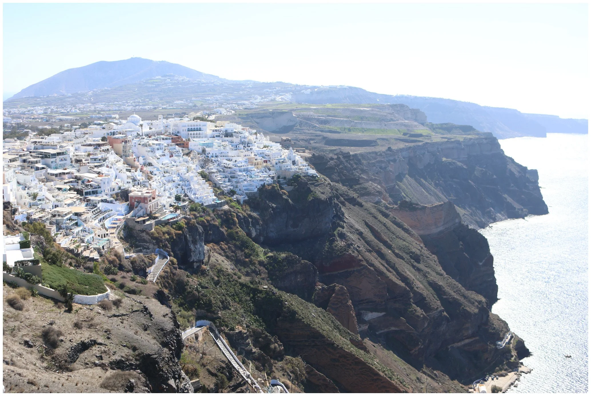 A coastal view of Santorini, Greece, with white buildings on steep cliffs overlooking the blue sea and a mountain in the background.