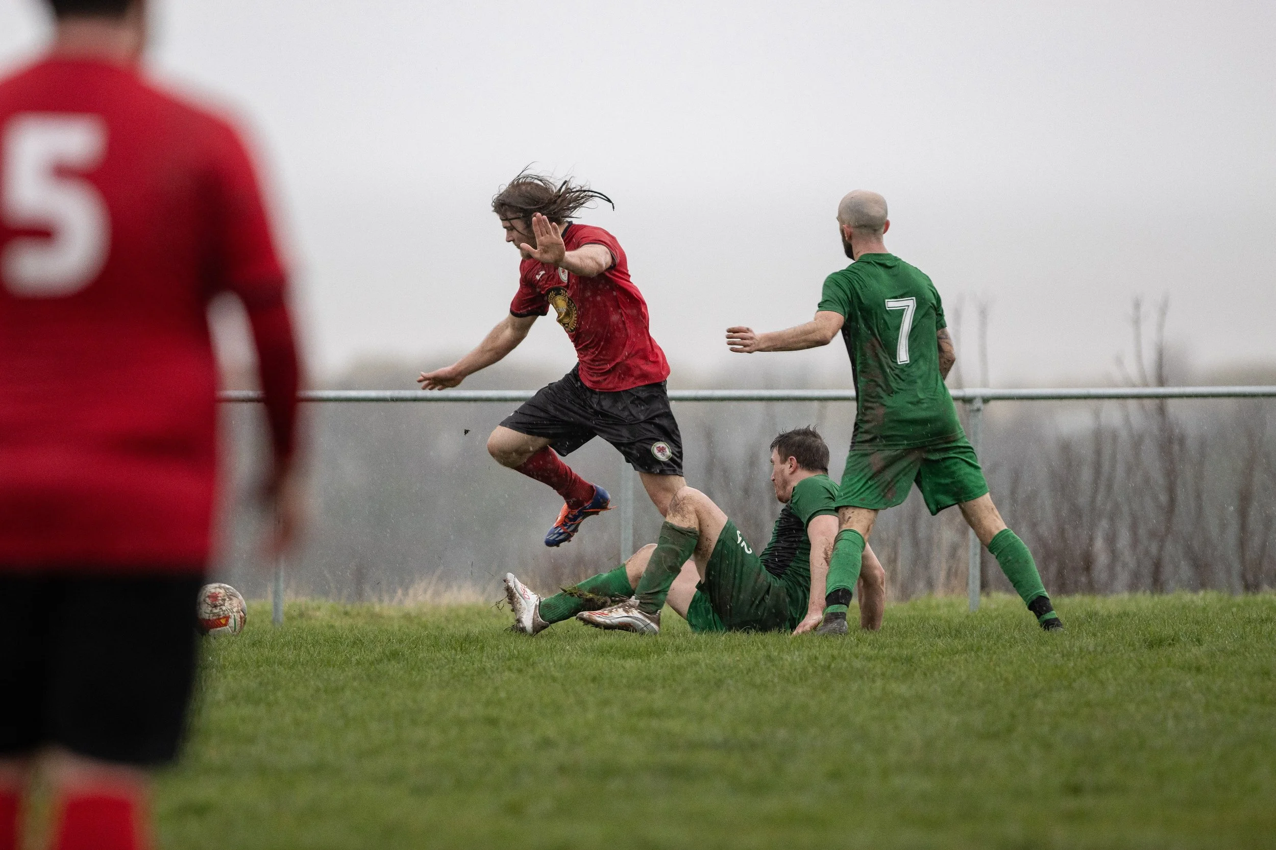 Soccer players in red and green uniforms competing for the ball on a wet field with a cloudy sky.