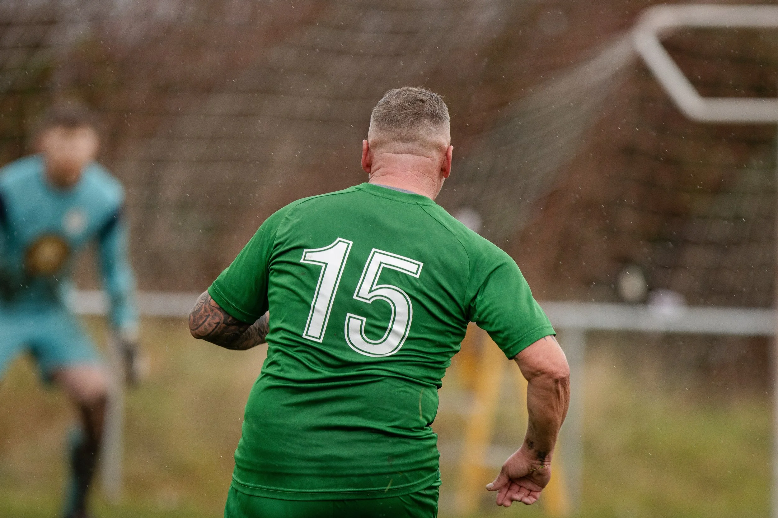 A soccer player wearing a green jersey with the number 15 on the back, running on a field during a game, with a goalkeeper in a teal kit in the background.