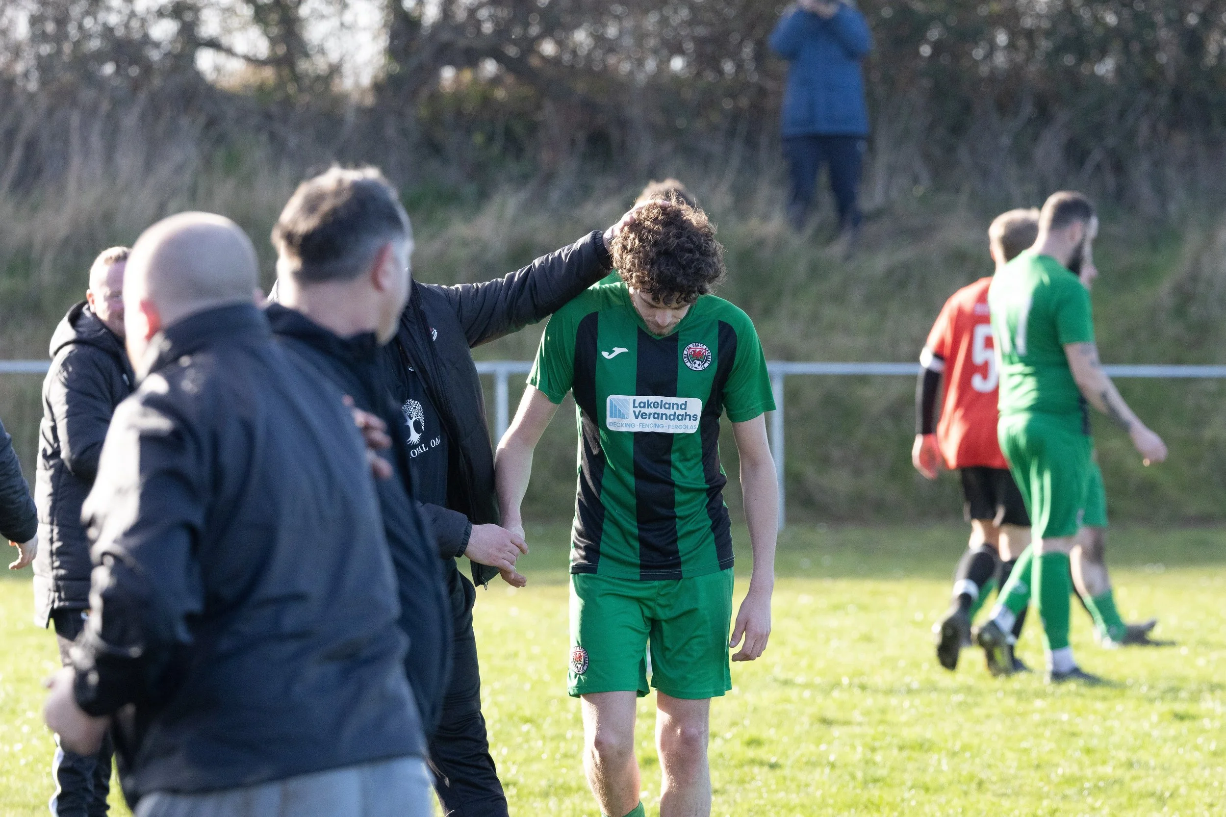 A soccer player in a green and black uniform is being comforted by a man with his hand on his head during a game, with other players and people in the background.