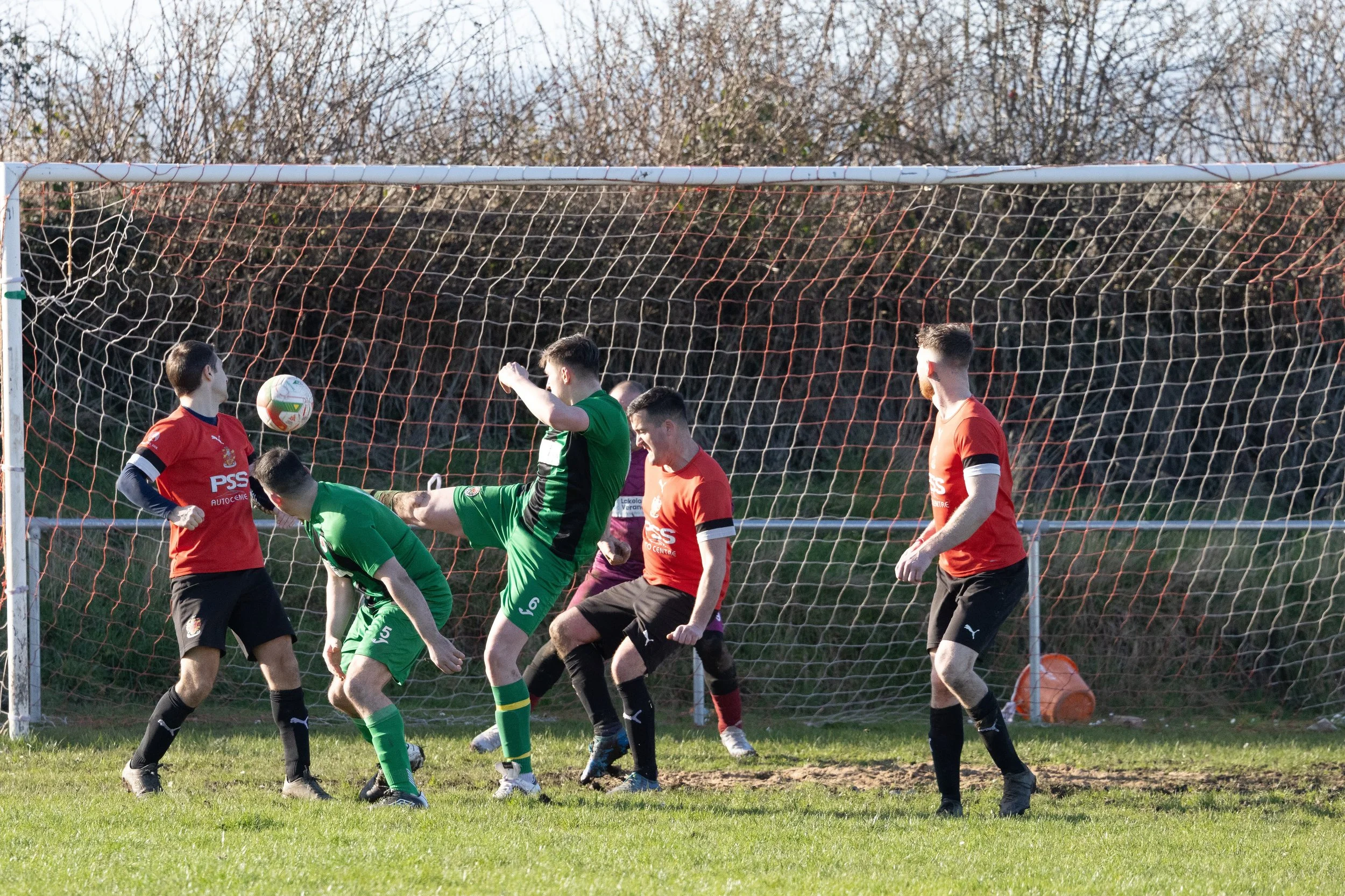 Group of young men playing soccer near the goal, with one kicking the ball as others watch on in a grass field.