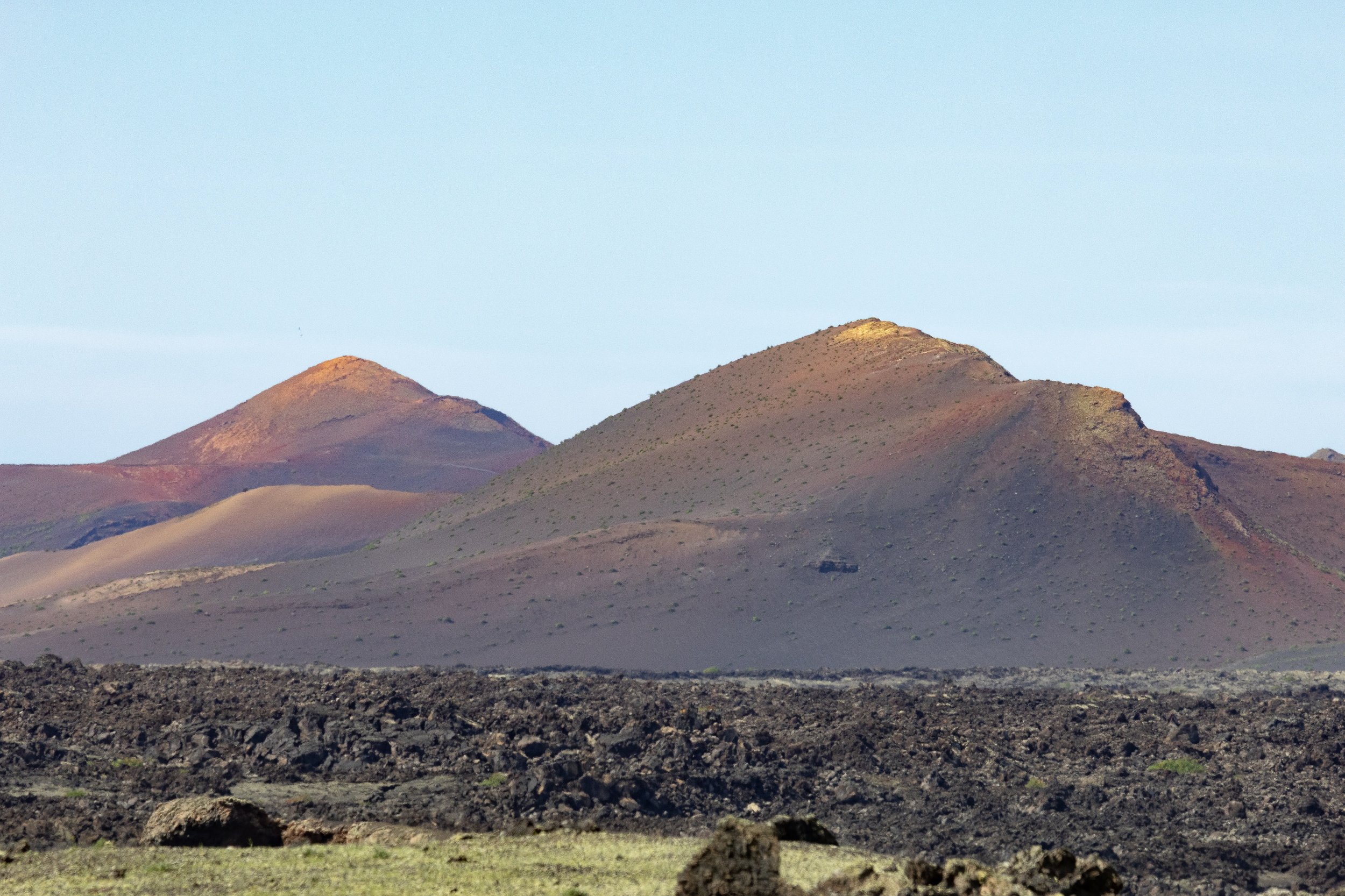 Multiple volcanic hills or mountains with dark and reddish-brown slopes, under a light blue sky.