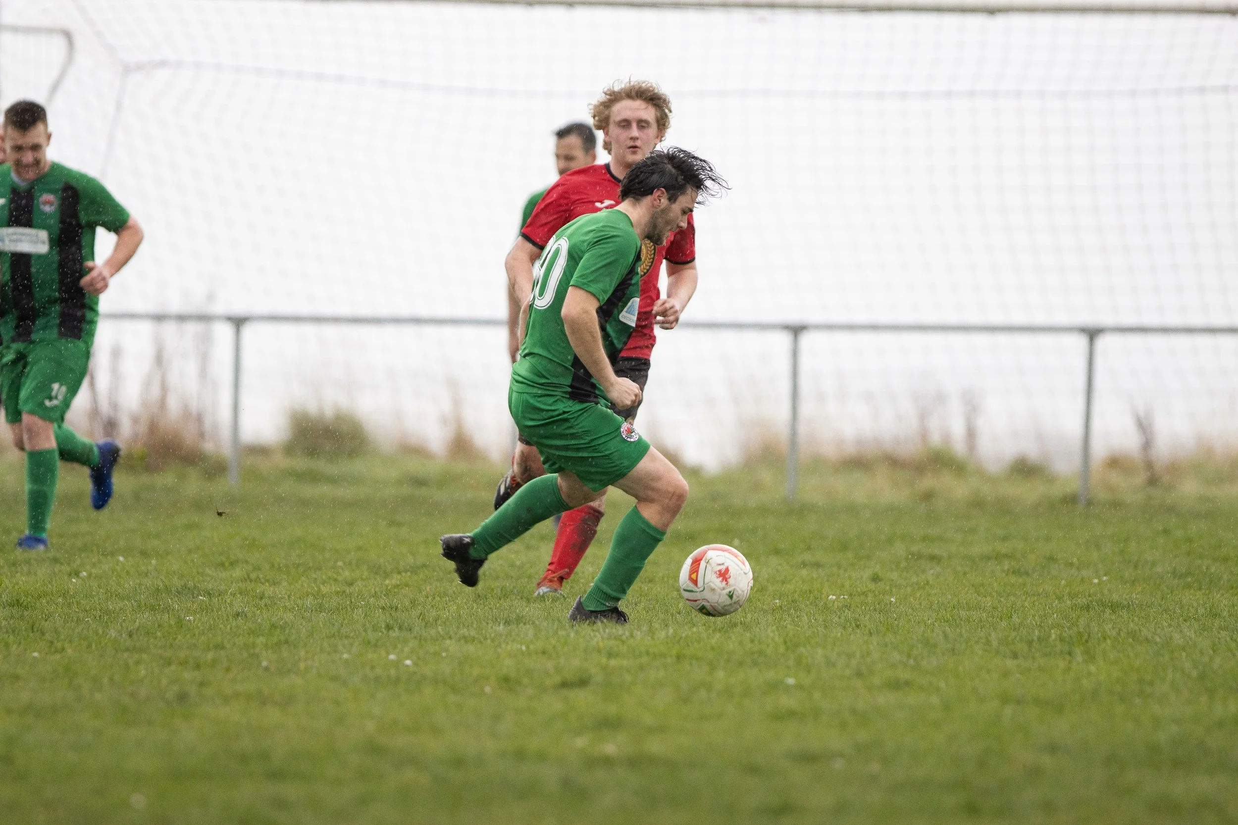 Soccer players competing for ball on field with goal net in background, one in green jersey and others in red and green jerseys.